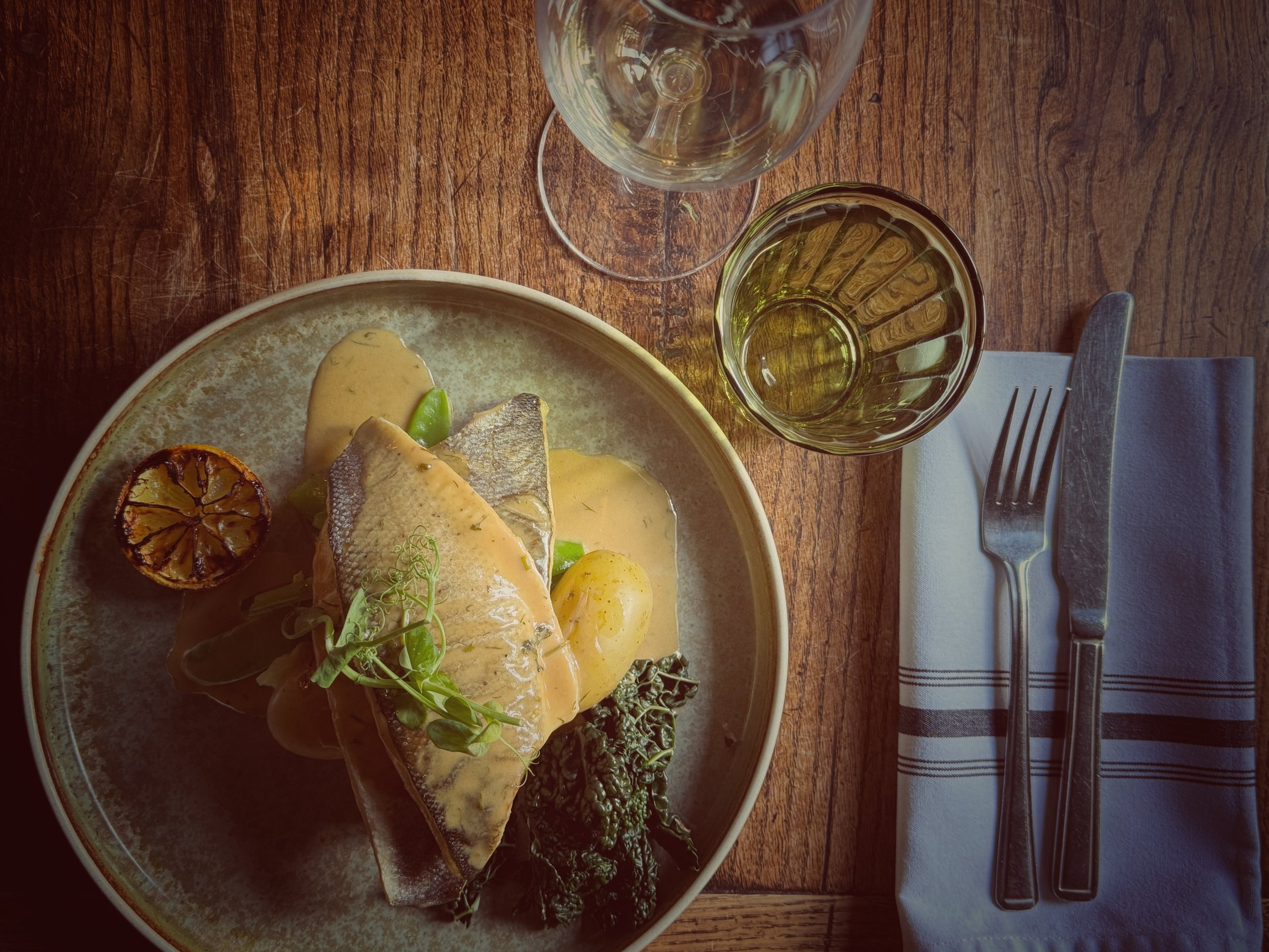 A plate of gourmet fish with lemon slices, grilled vegetables, and microgreens on a rustic wooden table, accompanied by a glass of white wine, a water glass, a fork, and a napkin with black and white stripes.