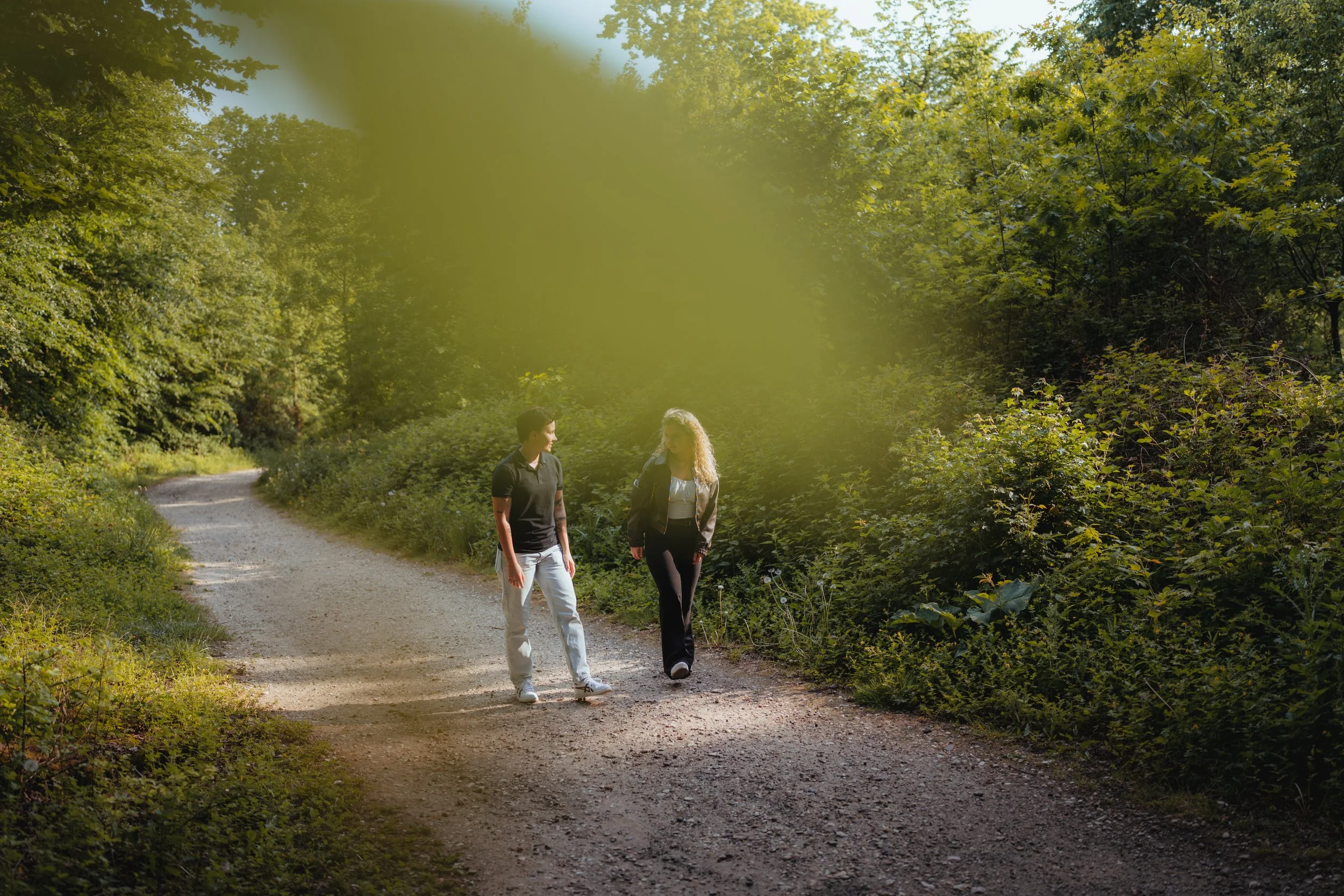 Zwei Frauen gehen zusammen auf einem Waldweg, umgeben von grünen Bäumen und Sträuchern, bei sonnigem Wetter und unterhalten sich.