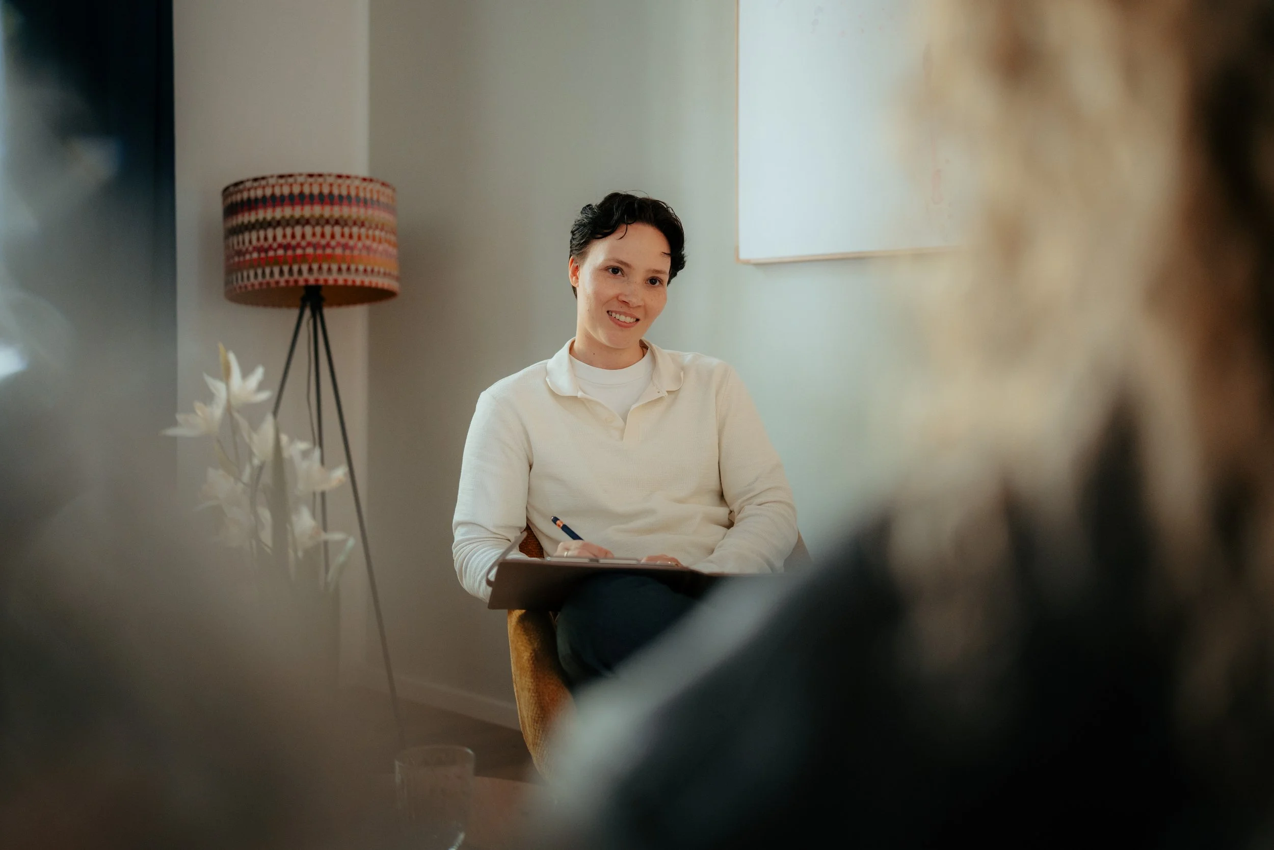 Eine junge Frau mit kurzen dunklen Haaren sitzt an einem Tisch, lächelt und hält einen Stift in der Hand. Im Hintergrund steht eine Stehlampe mit einem bunten Wollschirm und an der Wand hängt ein weißes Bild.