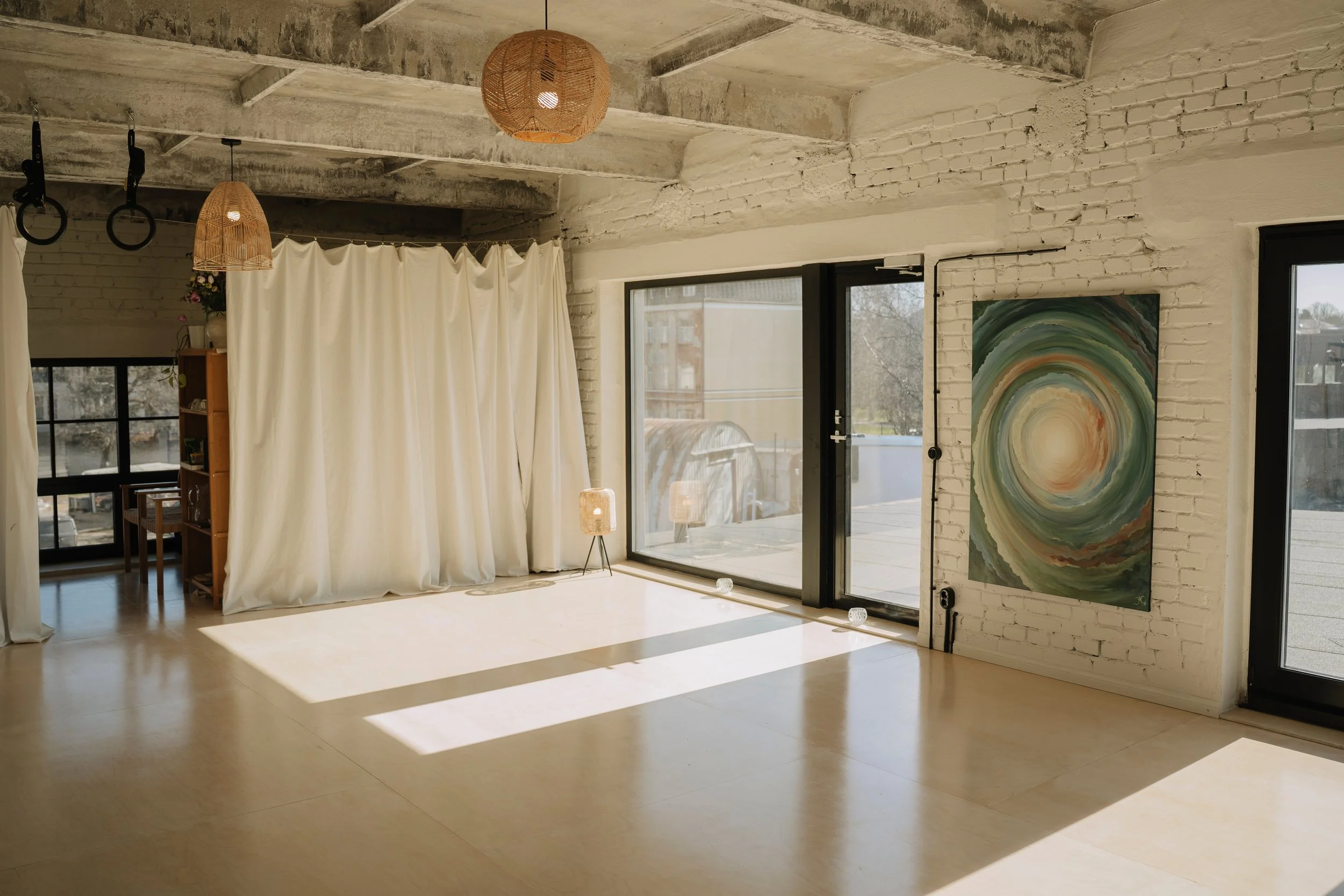 Bright, airy room with a white brick wall, large glass windows and doors, hanging wicker pendant lights, a colorful abstract painting on the wall, and a cream curtain partially covering an interior space.