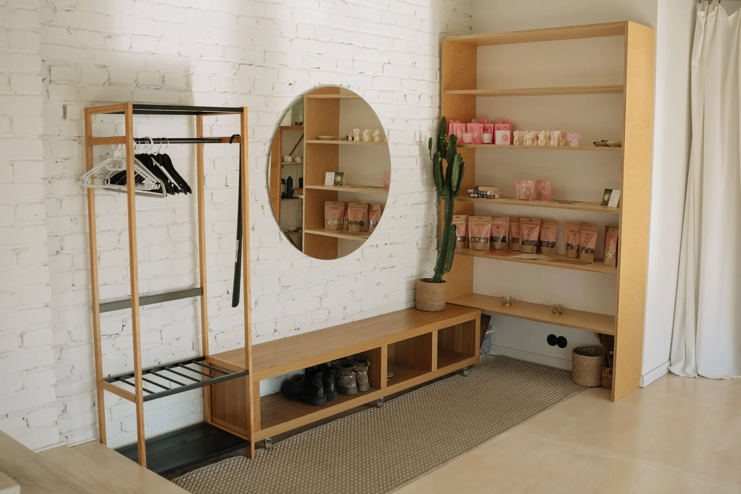 Interior of a boutique or a store with white brick walls, wooden shelving units, a mirror, and a plant. The shelves display pink and white packaged products and candles, with a valet stand holding black and white hangers and a few shoes underneath.