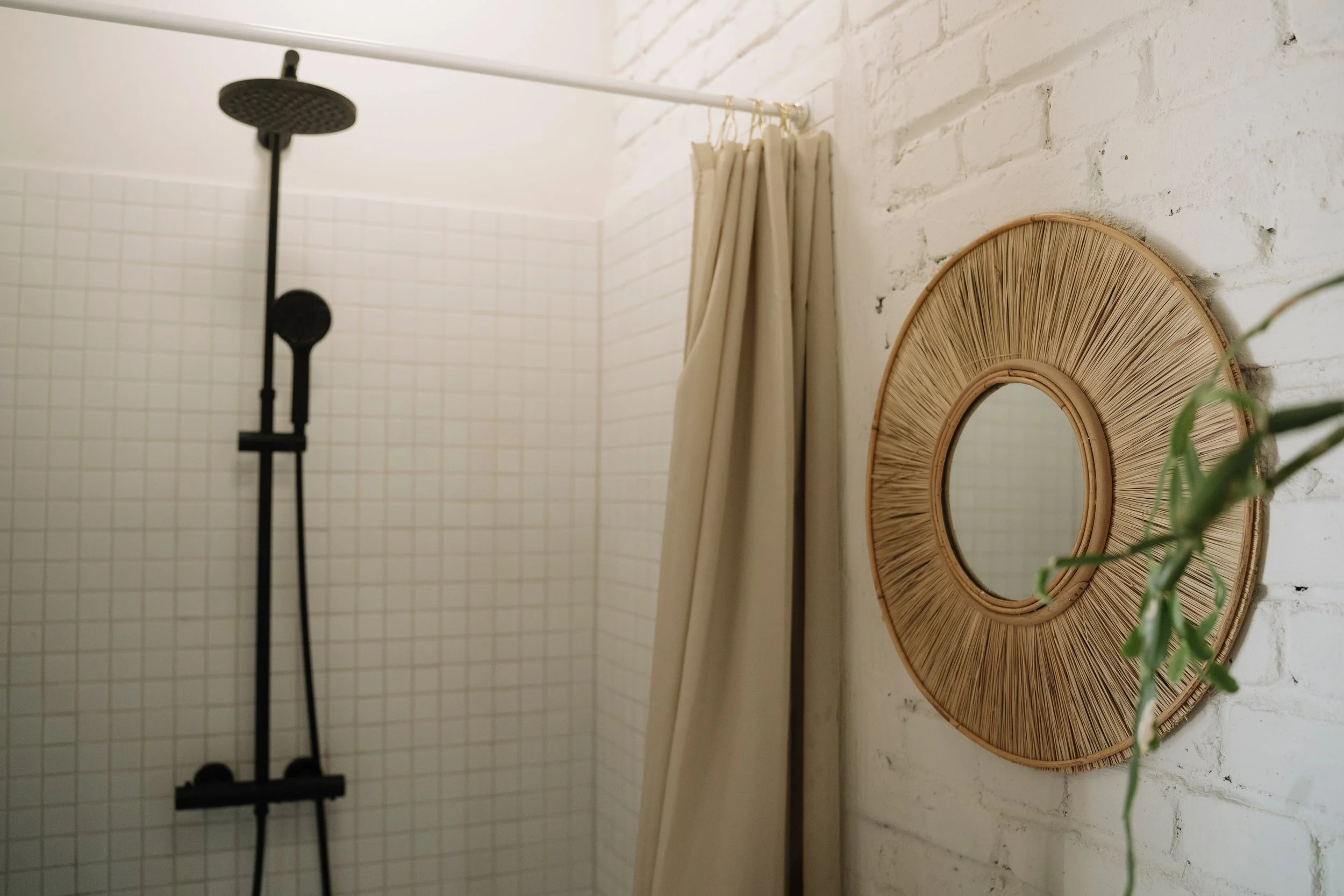 A bathroom shower with white tiled walls, a black showerhead and fixtures, a white curtain, a round wicker mirror, and a plant in the foreground.