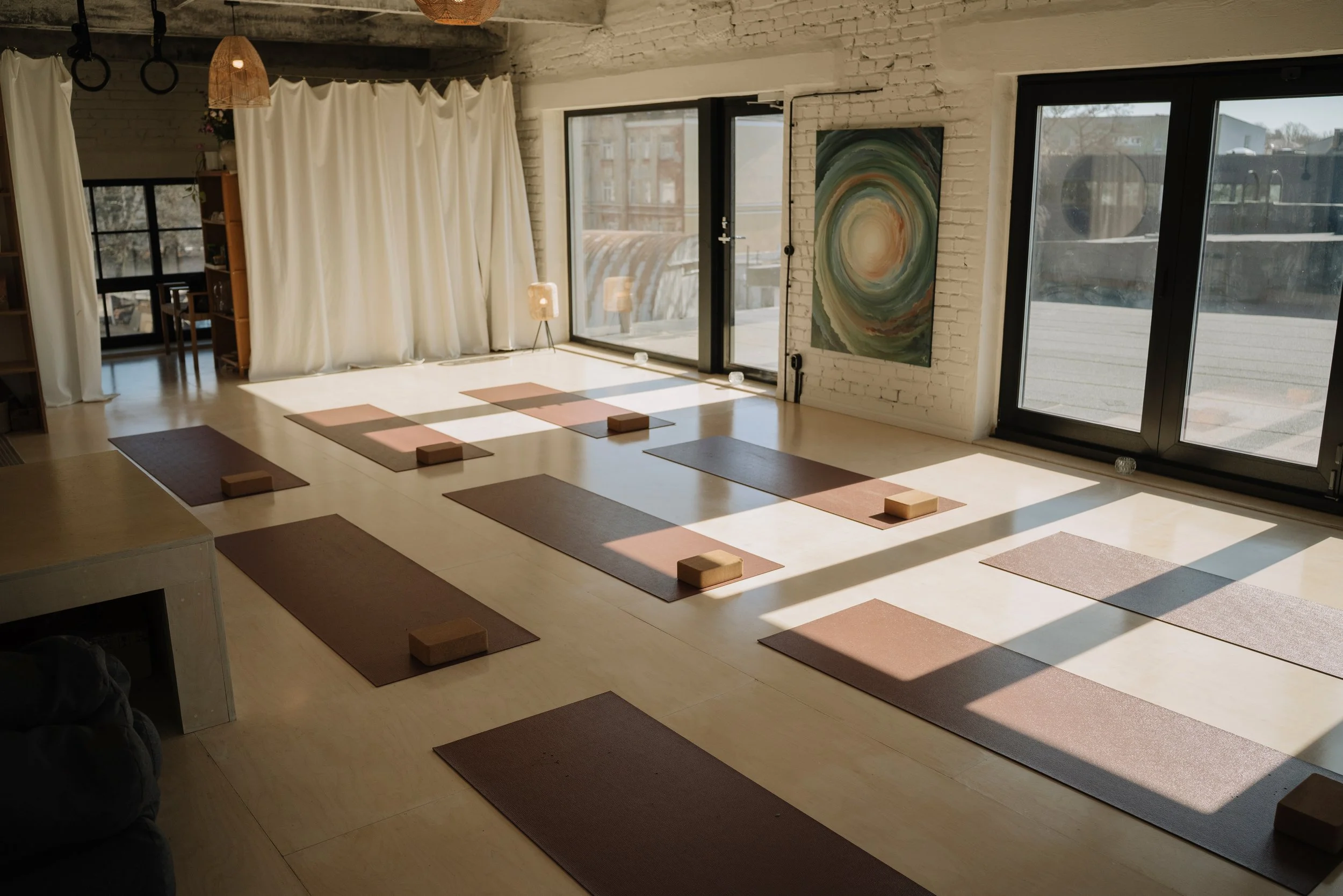 A bright yoga or meditation studio with sunlight streaming through large windows. The room has wooden floors, white brick walls, and a large colorful abstract painting. Yoga mats and blocks are set up on the floor for practice.