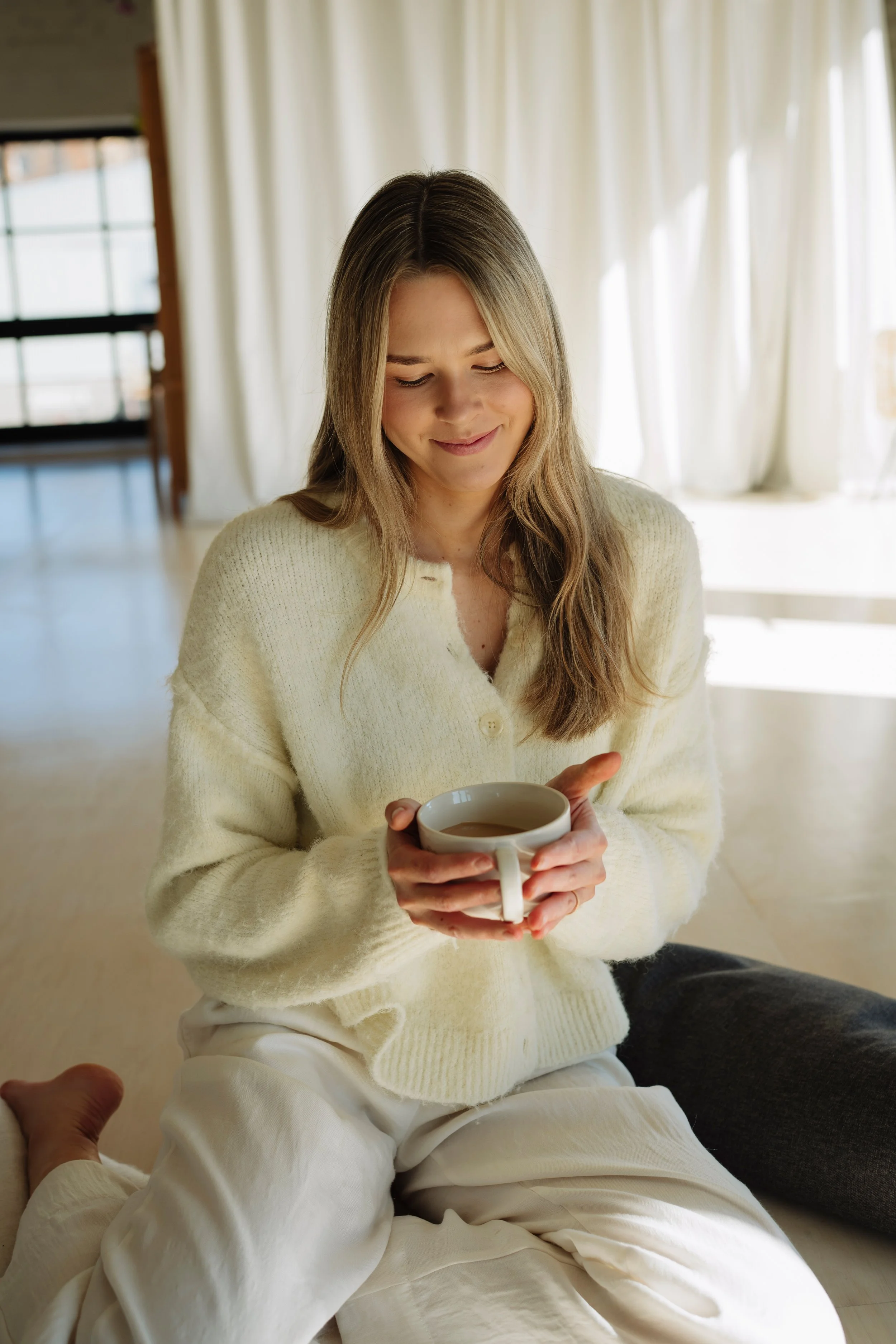 A woman with long blonde hair wearing a cream sweater and white pants, sitting on the floor and holding a mug of coffee, smiling softly while looking down, in a bright room with white curtains and wooden window frames.