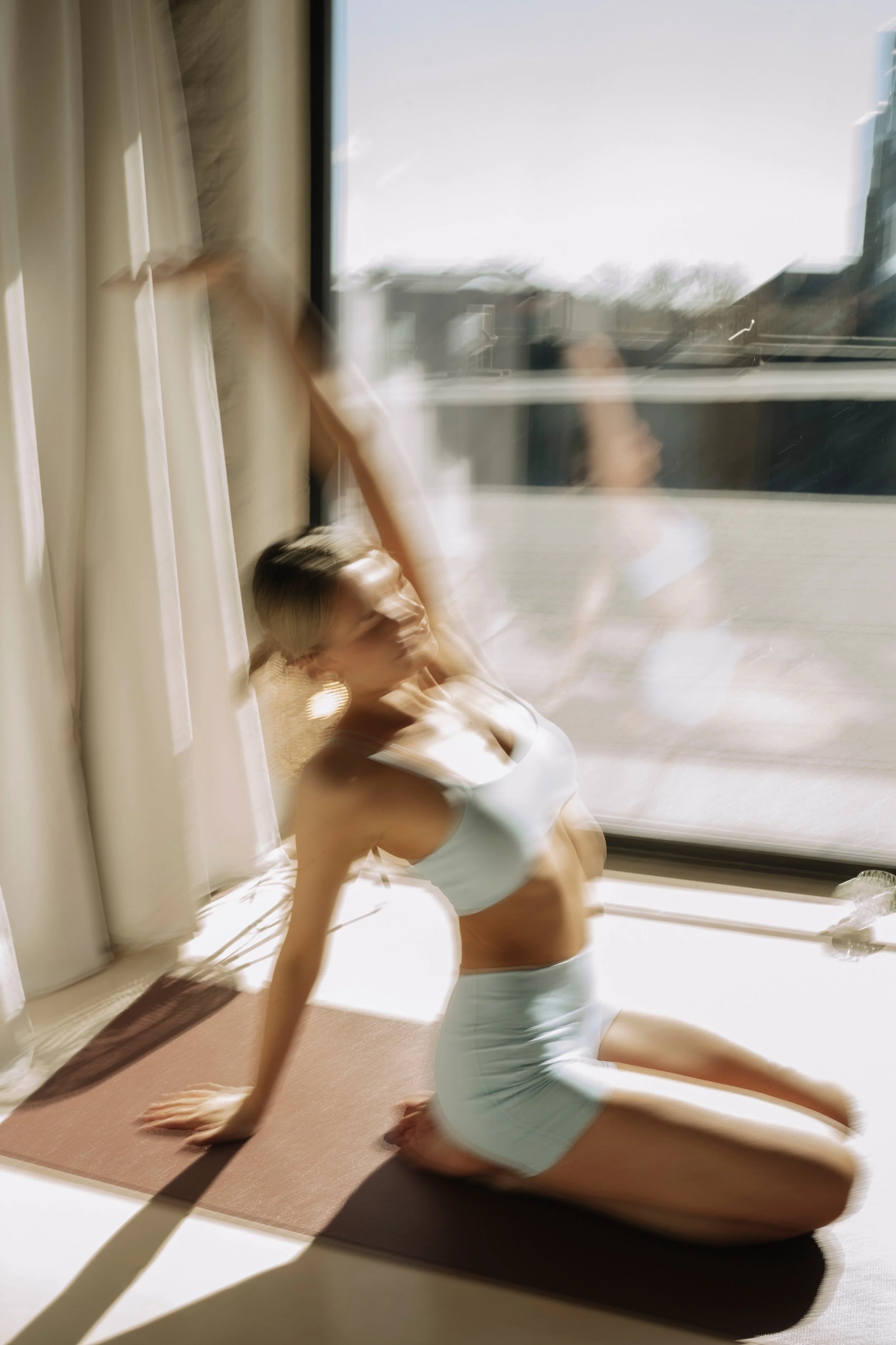 A woman in a white sports bra and shorts practicing yoga on a mat, kneeling on the floor in front of a window with curtains. The image appears blurred or distorted.