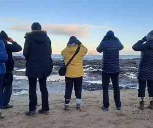 Group of five people standing on a beach facing the ocean, dressed in winter clothing.
