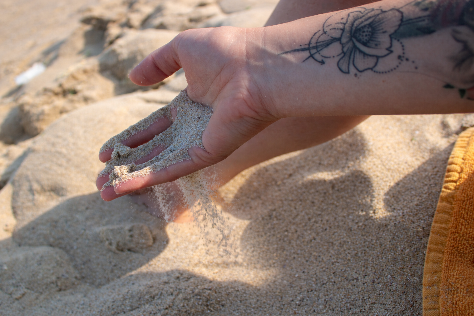 A person's hand with a floral tattoo on the forearm, holding and sifting sand on a beach.