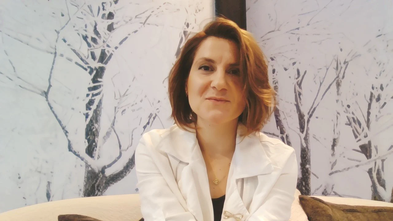 A woman with shoulder-length reddish-brown hair sitting on a beige sofa in front of a wall with a winter tree mural.