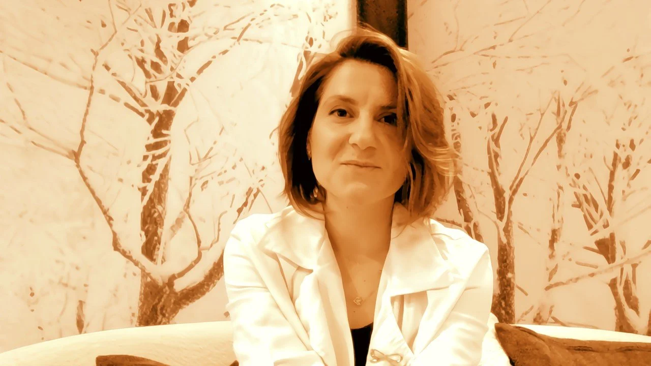 A woman with shoulder-length brown hair, smiling slightly, sitting in front of a mural of tree branches on the wall, wearing a white jacket.