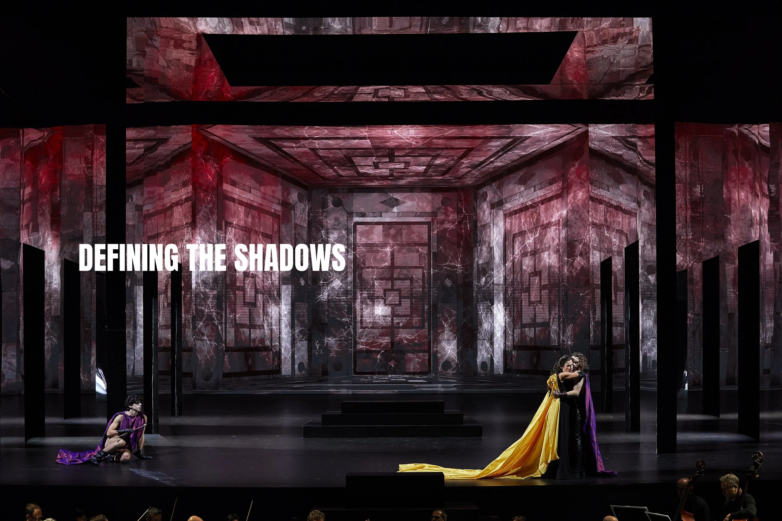 Stage scene from a theatrical performance with a modern set design featuring dark geometric patterns and lightning effects, three performers in colorful costumes, one sitting and two standing in an embrace, with the text "Defining the Shadows" displa