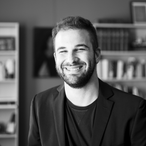 A smiling man with a beard and short hair, wearing a blazer, in an office or library setting with shelves of books in the background.