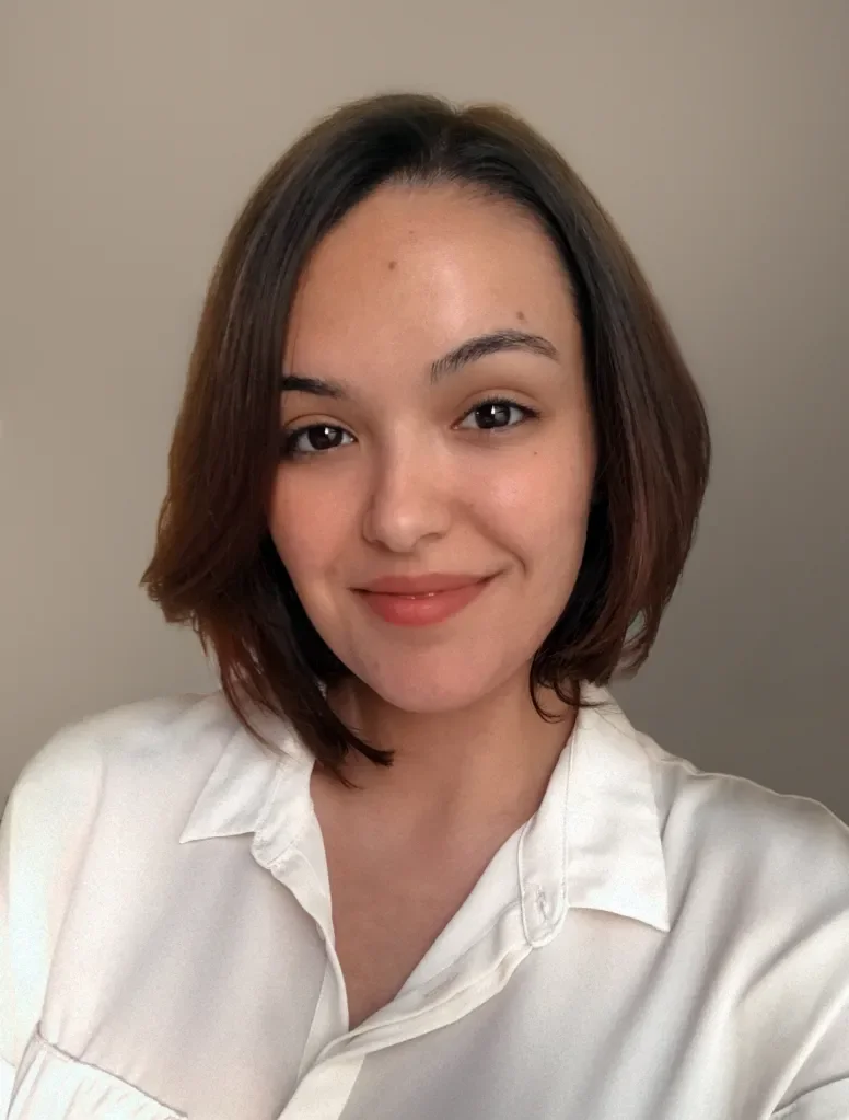 A young woman with shoulder-length brown hair and a natural smile, wearing a white shirt, standing against a plain, light-colored background.