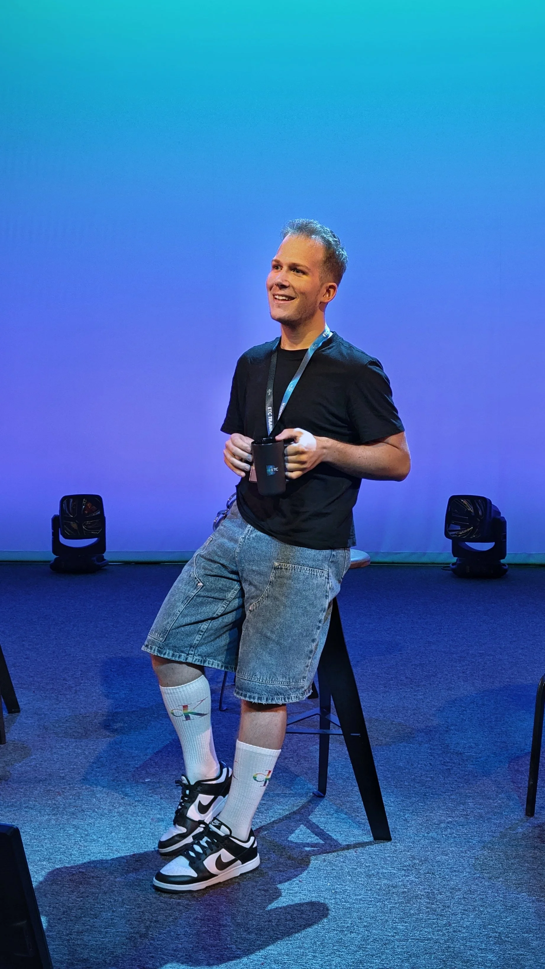 A man sitting on a tall stool on a stage, holding a mug, smiling, wearing a black T-shirt, gray shorts, white socks with Google logos, and black-and-white Nike sneakers, with stage lights in the background.