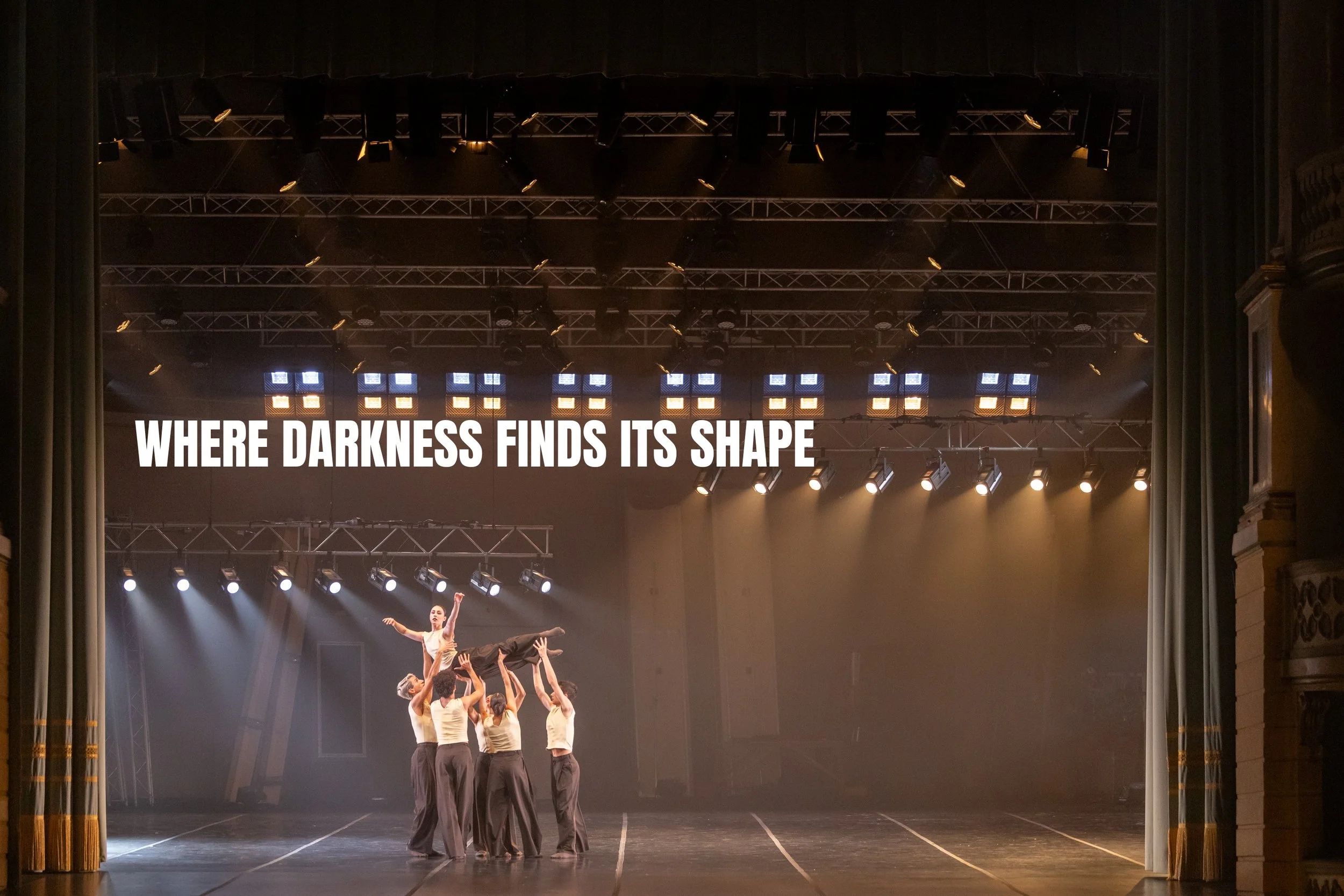 The image shows a stage with a group of dancers performing an act, with a male dancer being lifted by the others. Overhead stage lights illuminate the scene. There is a quote displayed on the stage backdrop that reads, "WHERE DARKNESS FINDS ITS SHAPE