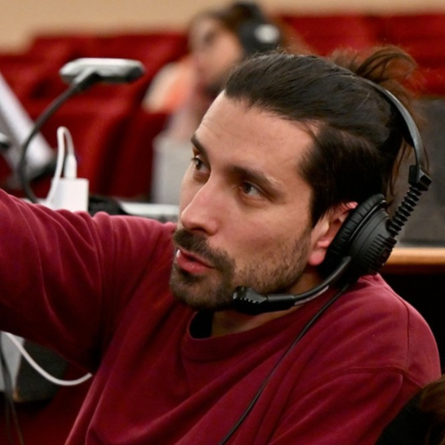 A man with dark hair and a beard wearing a maroon shirt and black headphones, speaking into a microphone in a recording studio or radio station.