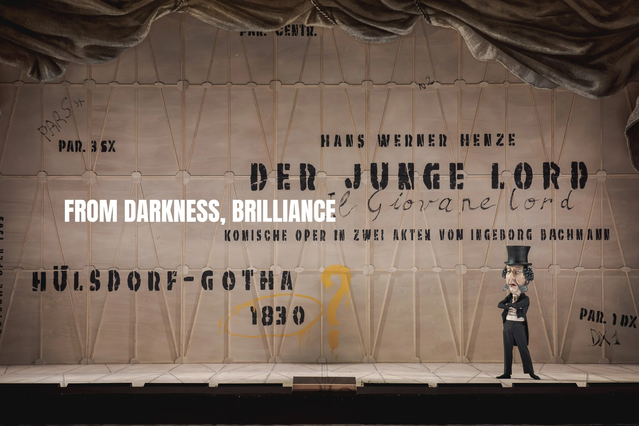 A theatrical stage backdrop with various historical references and German words. There is a puppet or character with a top hat standing on the stage. The words 'From Darkness, Brilliance' are overlaid in white text.