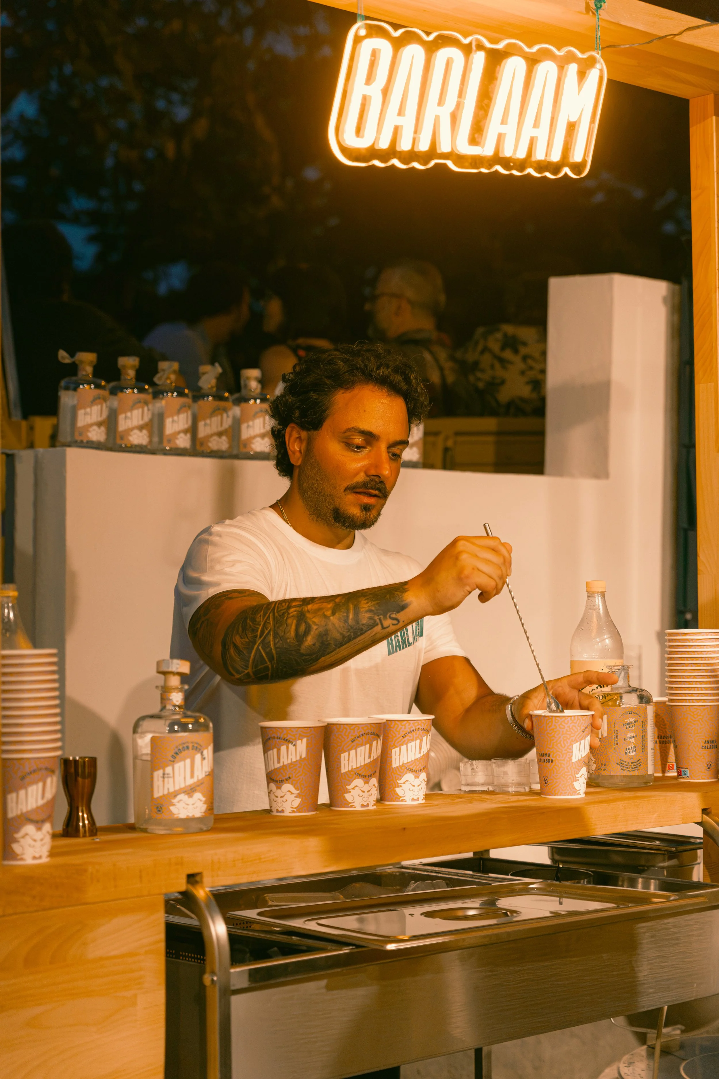 Un uomo con tatuaggi alla braccia lavora in uno stand con bicchieri e bottiglie di bibite alla birra, sotto un'insegna luminosa con la scritta 'BARLHAM'.