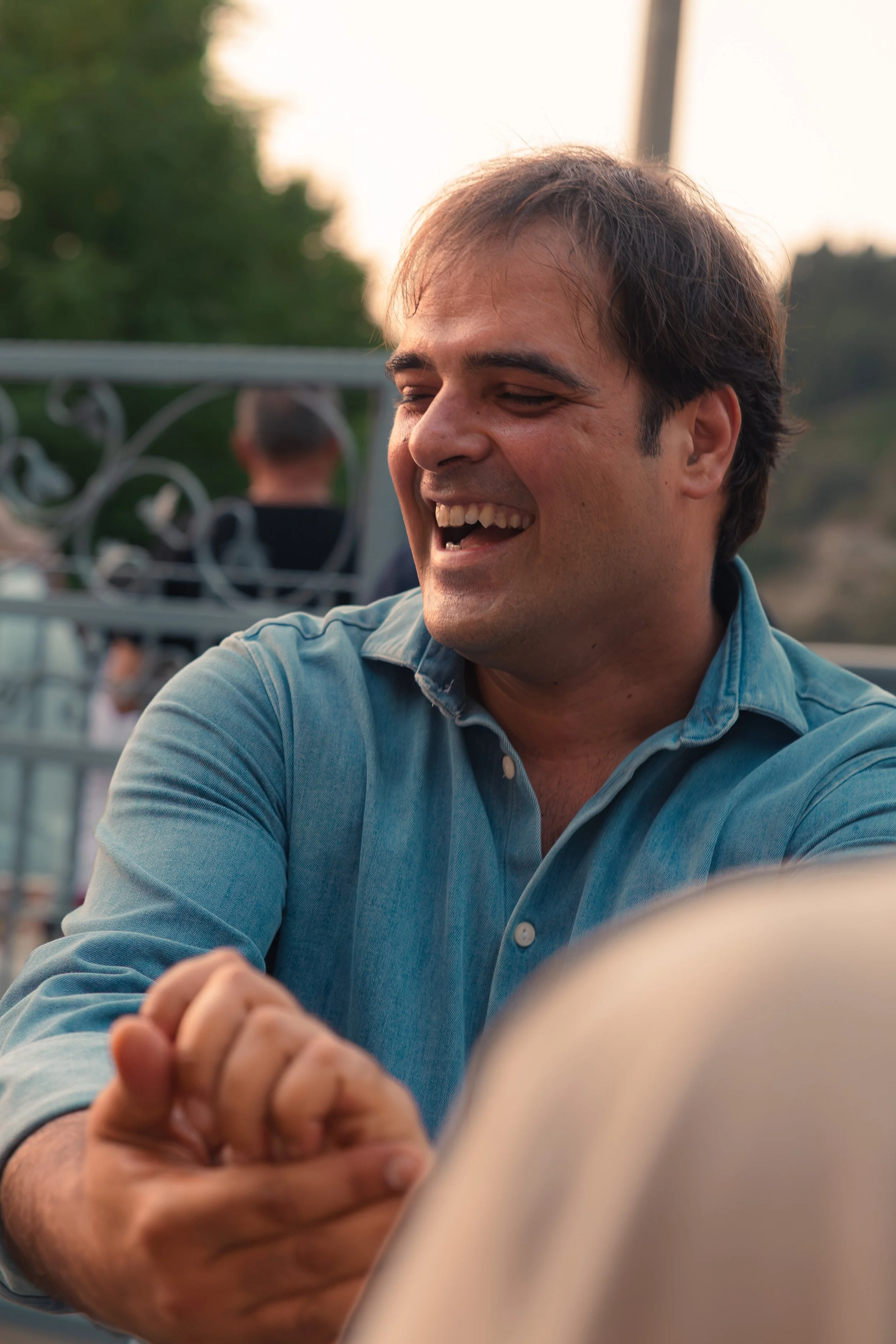 Uomo con sorriso che indossa una camicia azzurra, seduto all'aperto durante il tramonto
