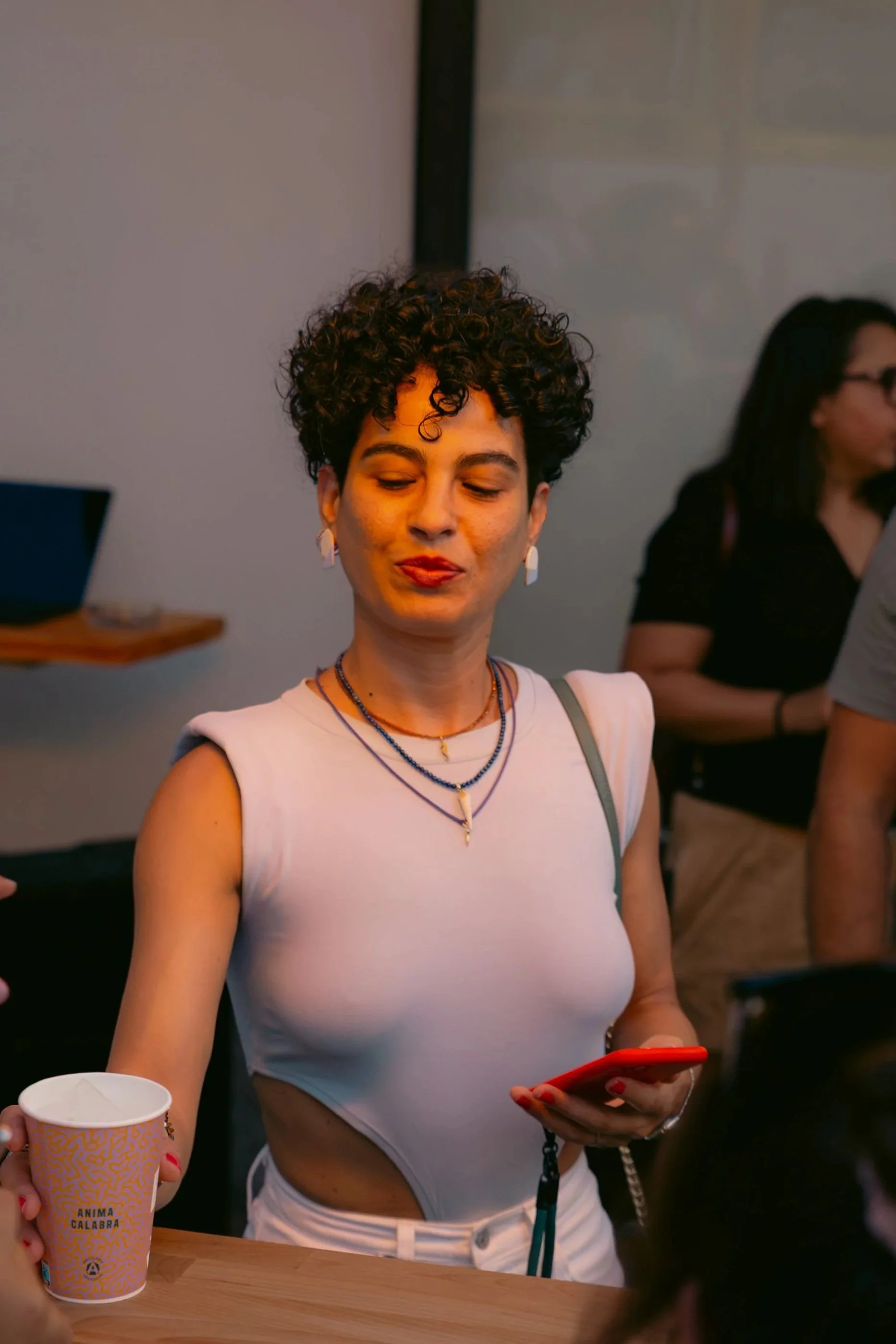 Donna con capelli riccioli corti e pelle chiara che guarda il suo telefono e tiene una tazza di carta con scritte in italiano. È vestita con top bianco e sviluppato da collane.