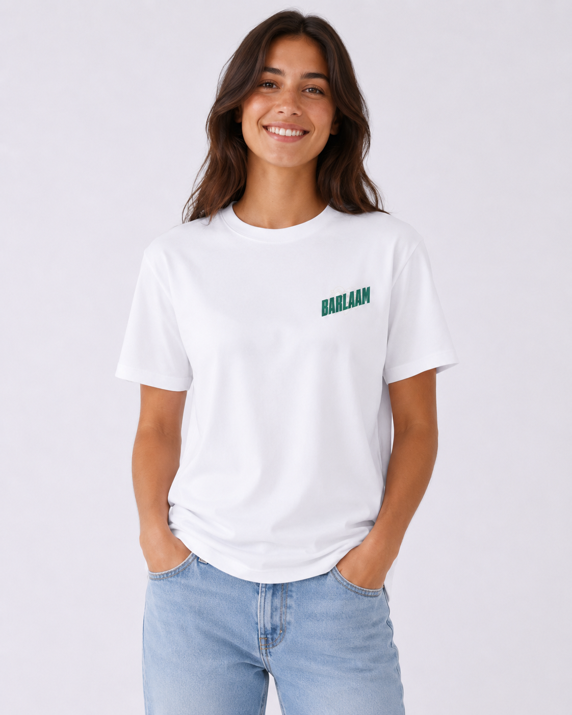 Una donna con capelli castani, sorriso e abbigliamento casual che indossa una maglietta bianca con la scritta "BARLAAM" di colore verde e jeans blu, in piedi davanti a uno sfondo bianco.