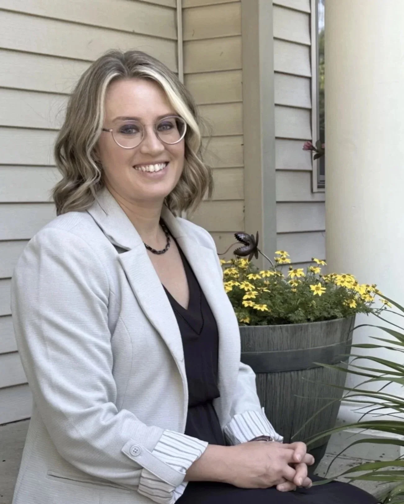 A woman with blonde, wavy hair and glasses sitting outdoors in front of a beige house wall with yellow flowers in a gray pot.