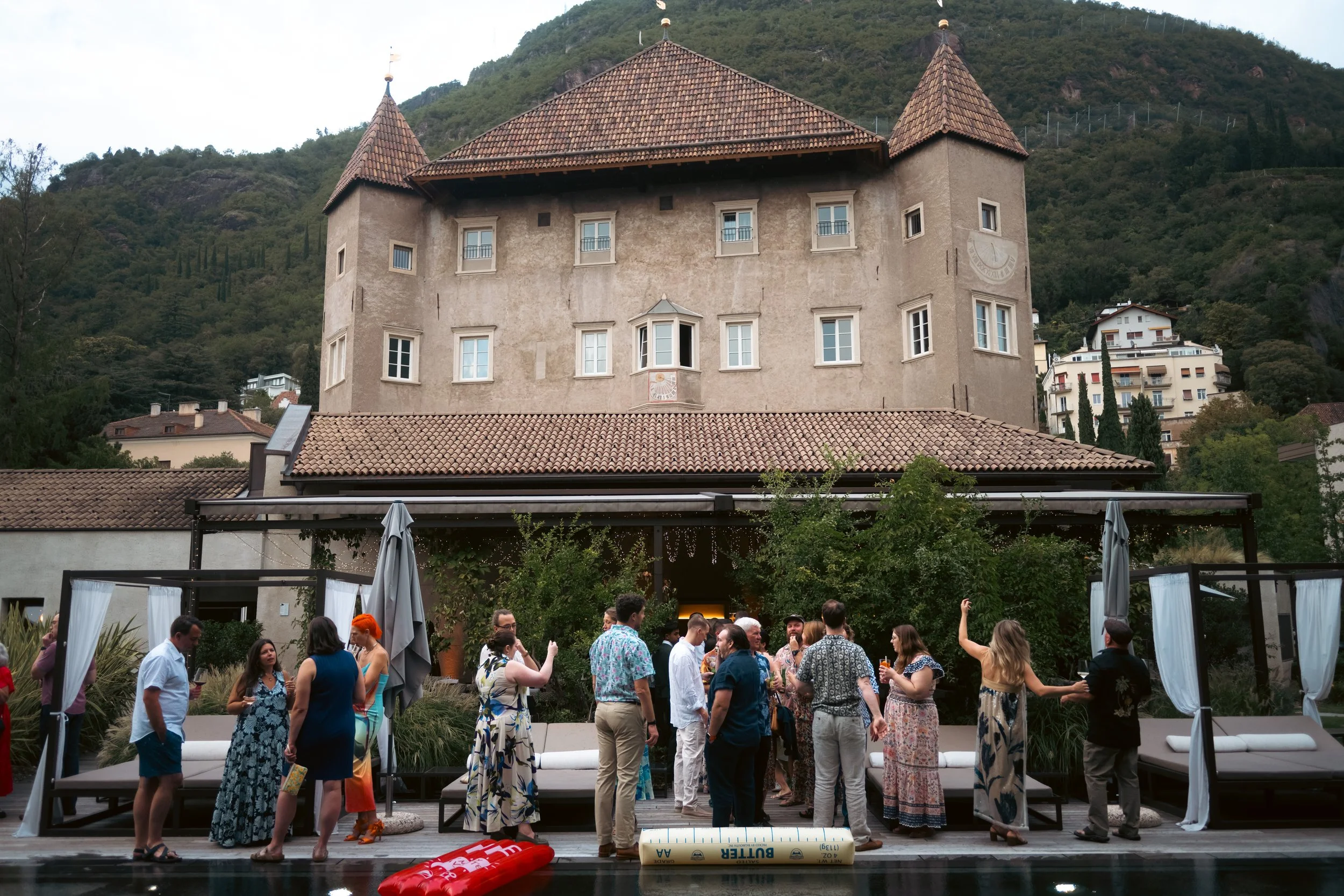  Wedding guests socializing outside Castel Hörtenberg with the Italian Dolomites in the background during a luxury destination welcome party. 