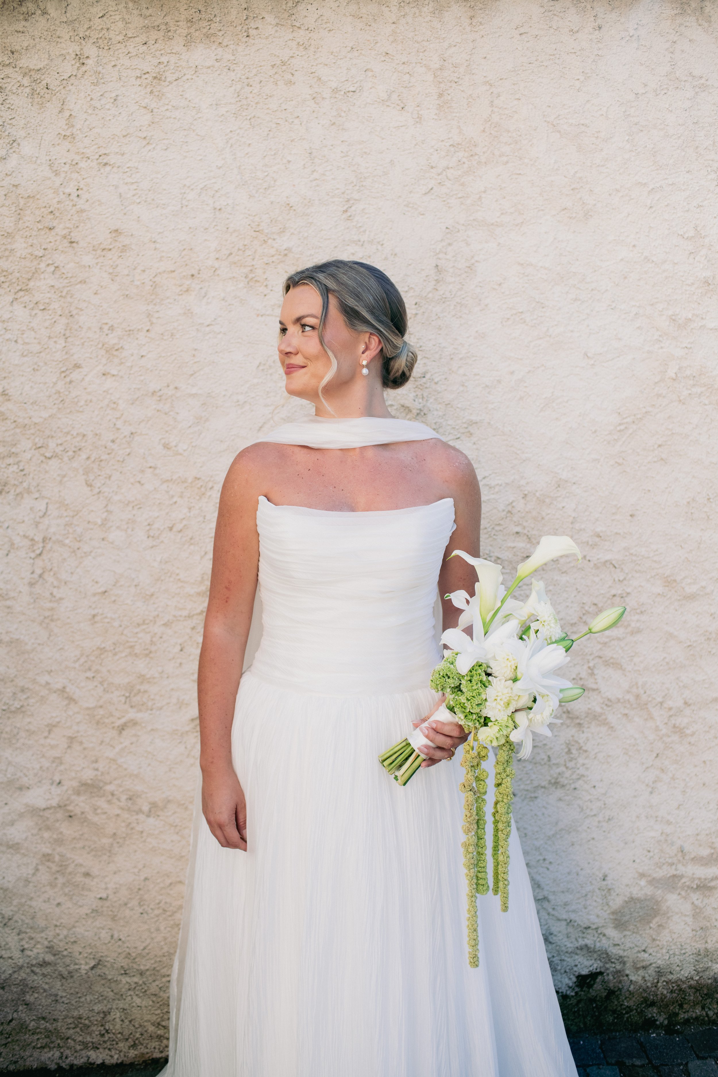 A woman dressed in a white wedding gown holding a bouquet of white lilies and green flowers, standing against a beige textured wall.