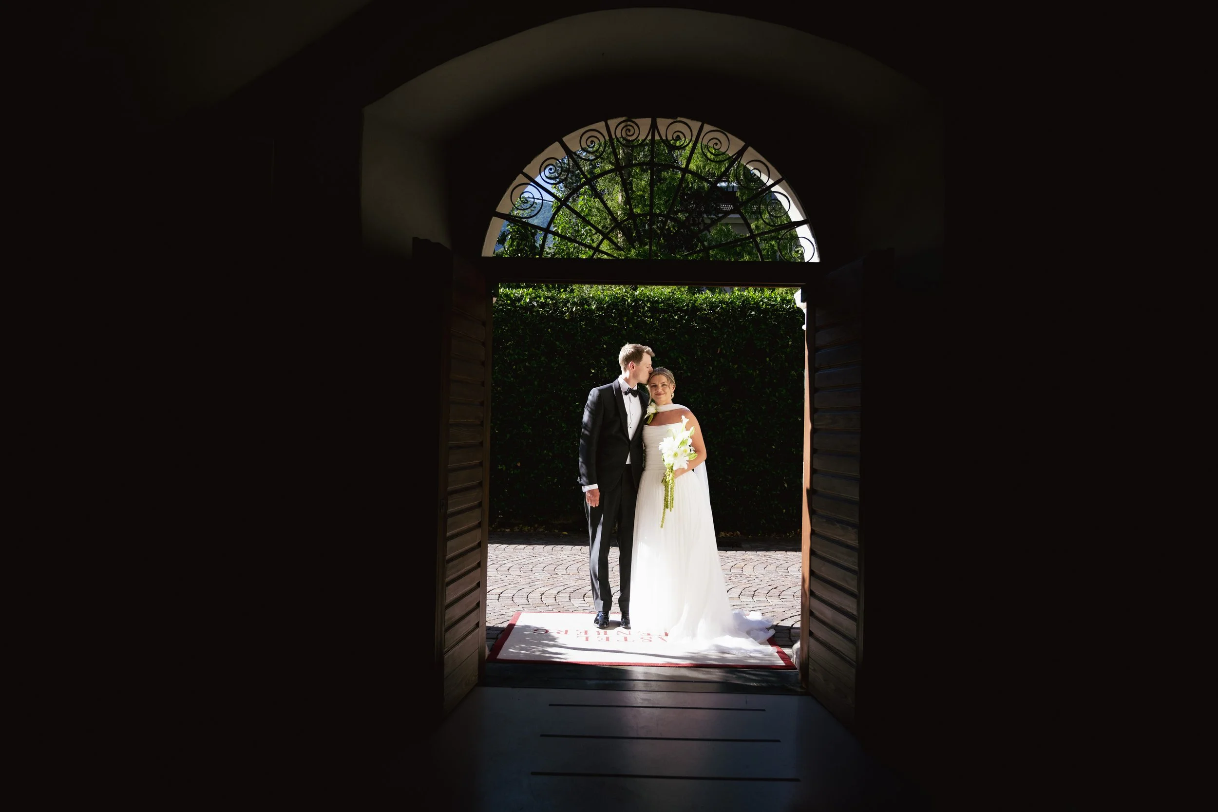 A newlywed couple stands in a doorway with open shutters, backlit by sunlight, with trees and greenery outside. The groom wears a black tuxedo with a bow tie, and the bride is in a white wedding dress, holding a bouquet of white flowers. They are looking at each other.