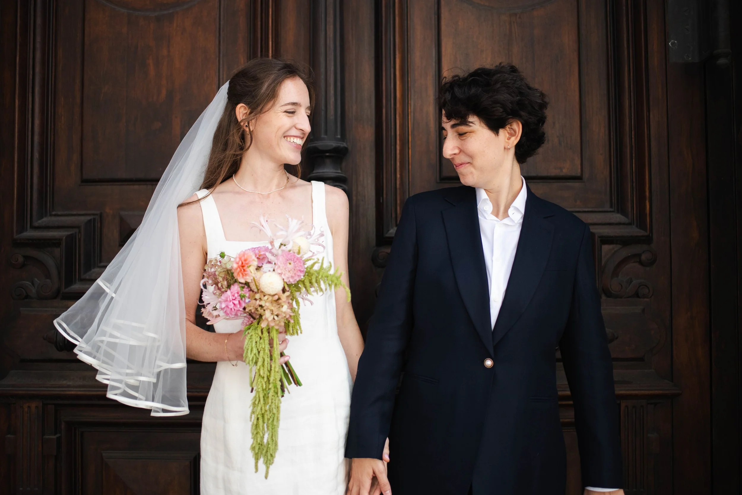 Two women, one in a white wedding dress holding a bouquet of flowers and the other in a dark suit, stand close together in front of a wooden backdrop, smiling and holding hands.