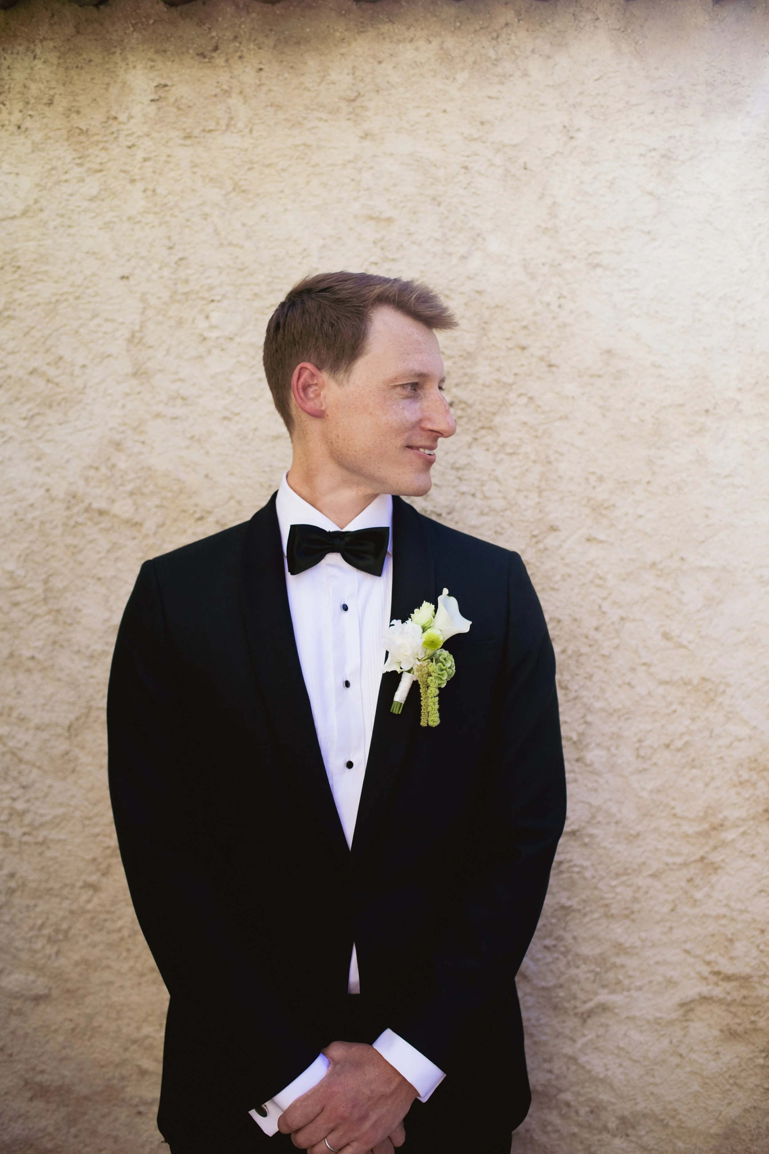 A groom in a tuxedo with a boutonniere stands against a textured beige wall, smiling and looking to the side.