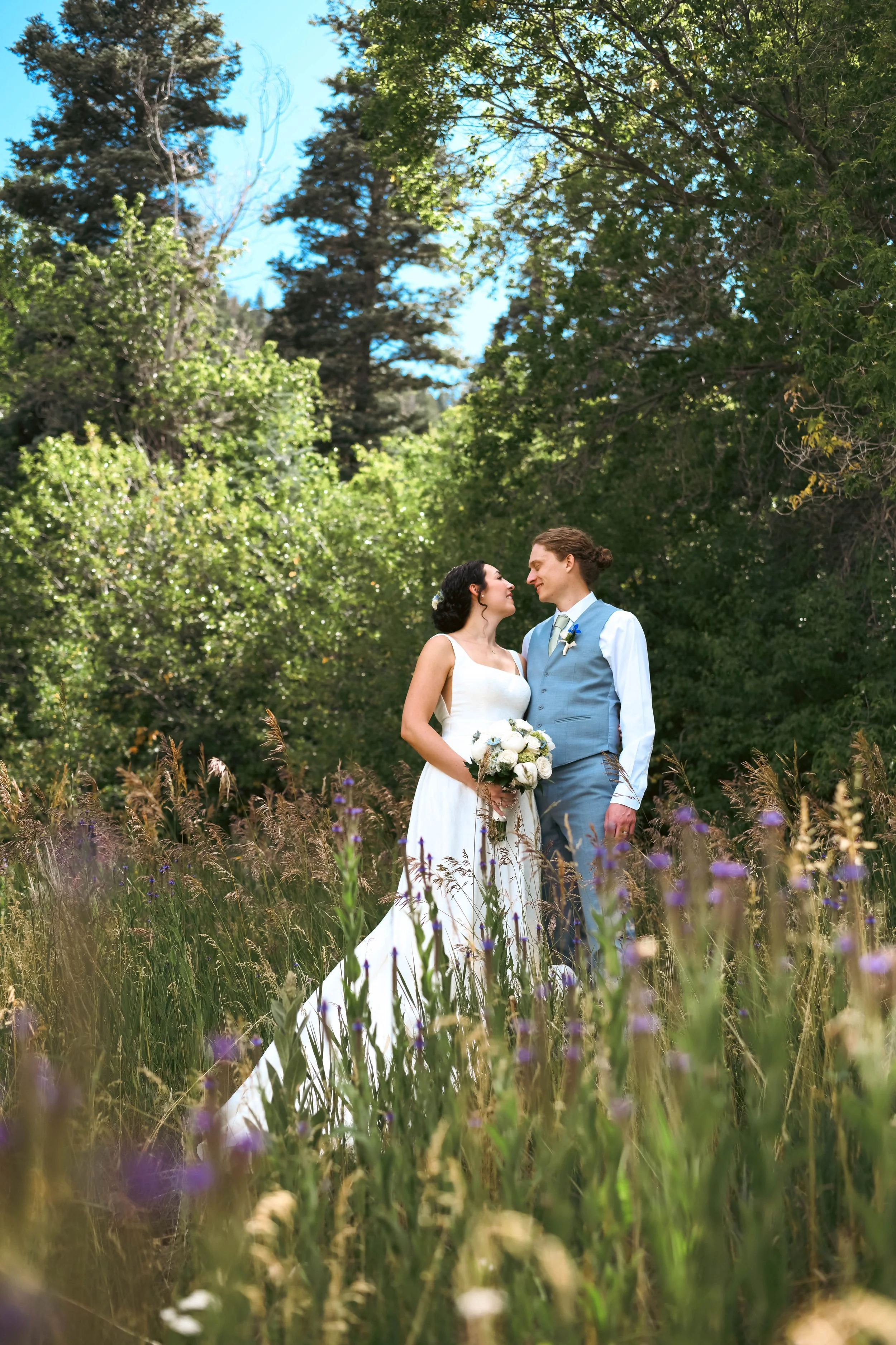 A bride and groom stand close in a lush New Mexico setting, surrounded by tall grass and wildflowers, with dense green trees behind them and a clear blue sky above. The bride holds a bouquet of white roses, and they look at each other lovingly.