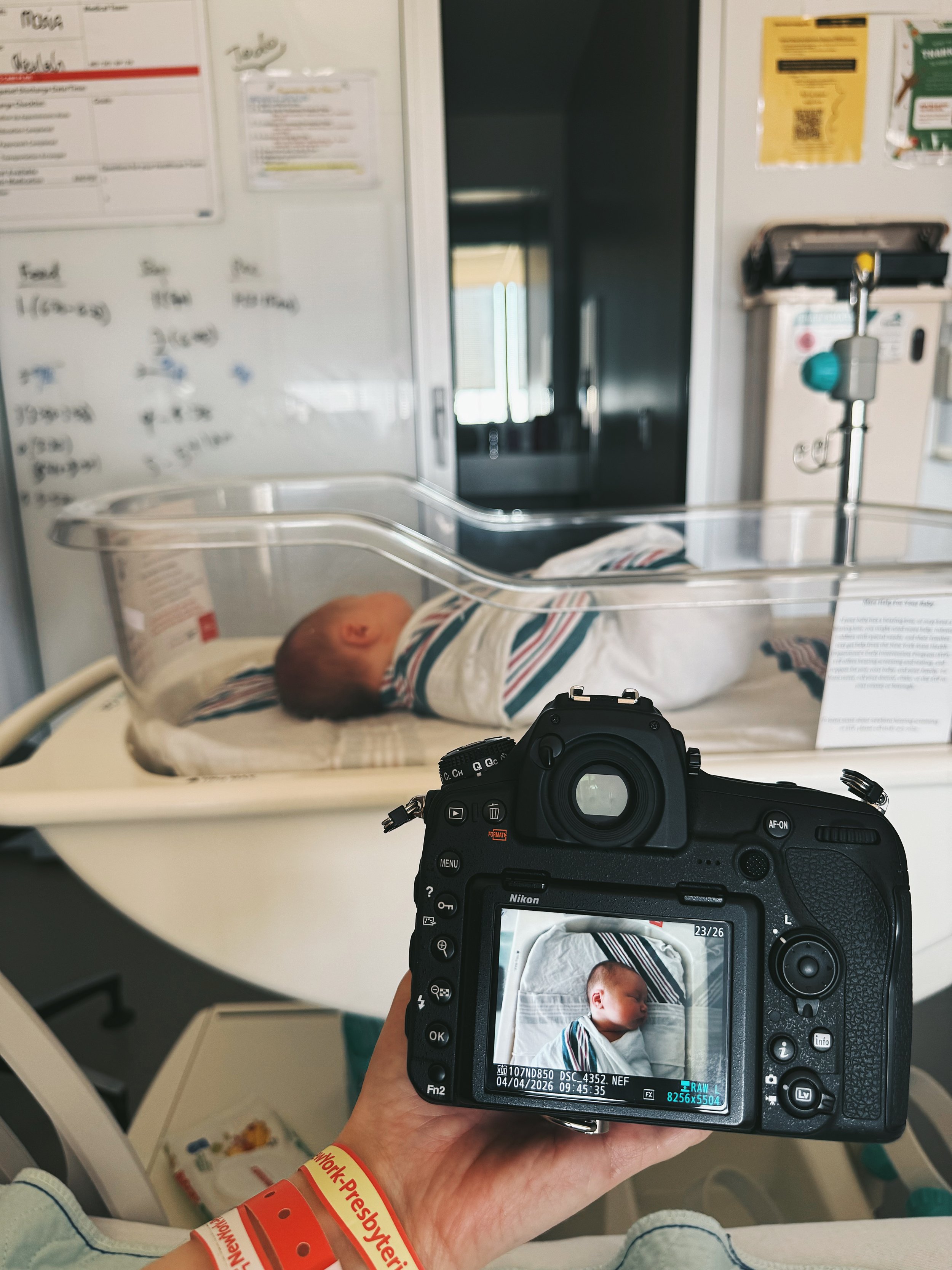 A photographer holds a camera displaying a crisp, high-resolution image of a newborn baby in a hospital setting, capturing the first moments of life through a professional lens.
