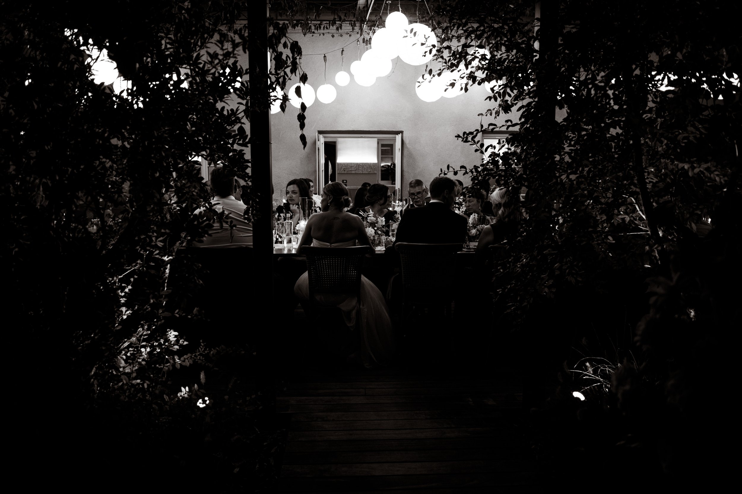 A black and white photo of a dining event seen through a natural opening in bushes. The image shows a guest table with people seated, illuminated by hanging lights above. There is a doorway in the background with large mirrors reflecting the room.
