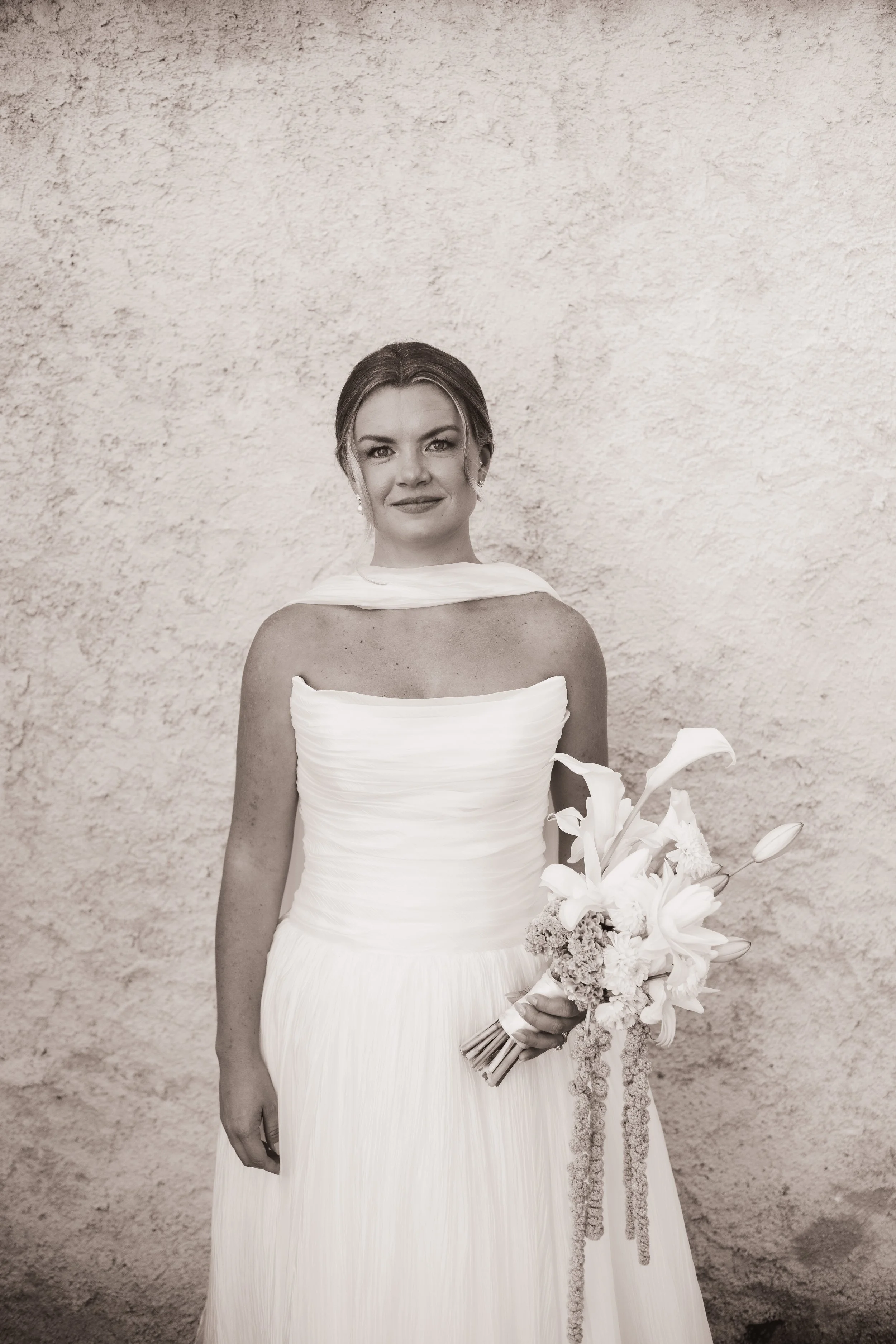 A woman in a white wedding dress holding a bouquet of flowers, standing in front of a textured wall.