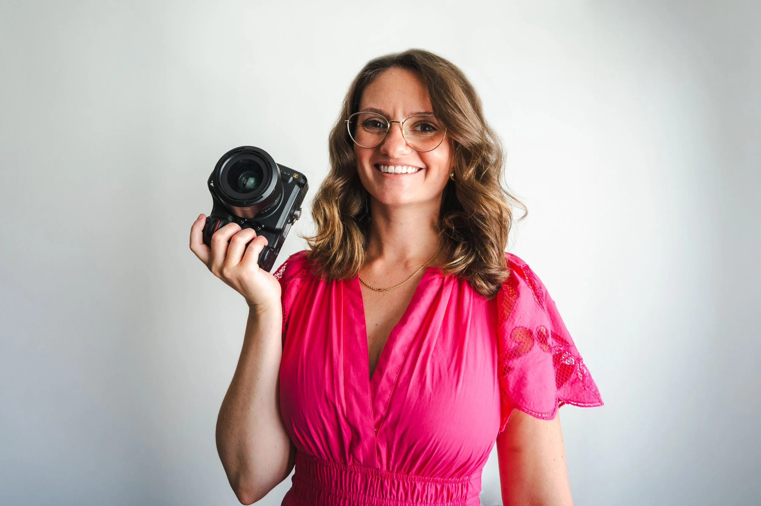 A woman with wavy brown hair wearing glasses and a pink dress, smiling and holding a professional camera.
