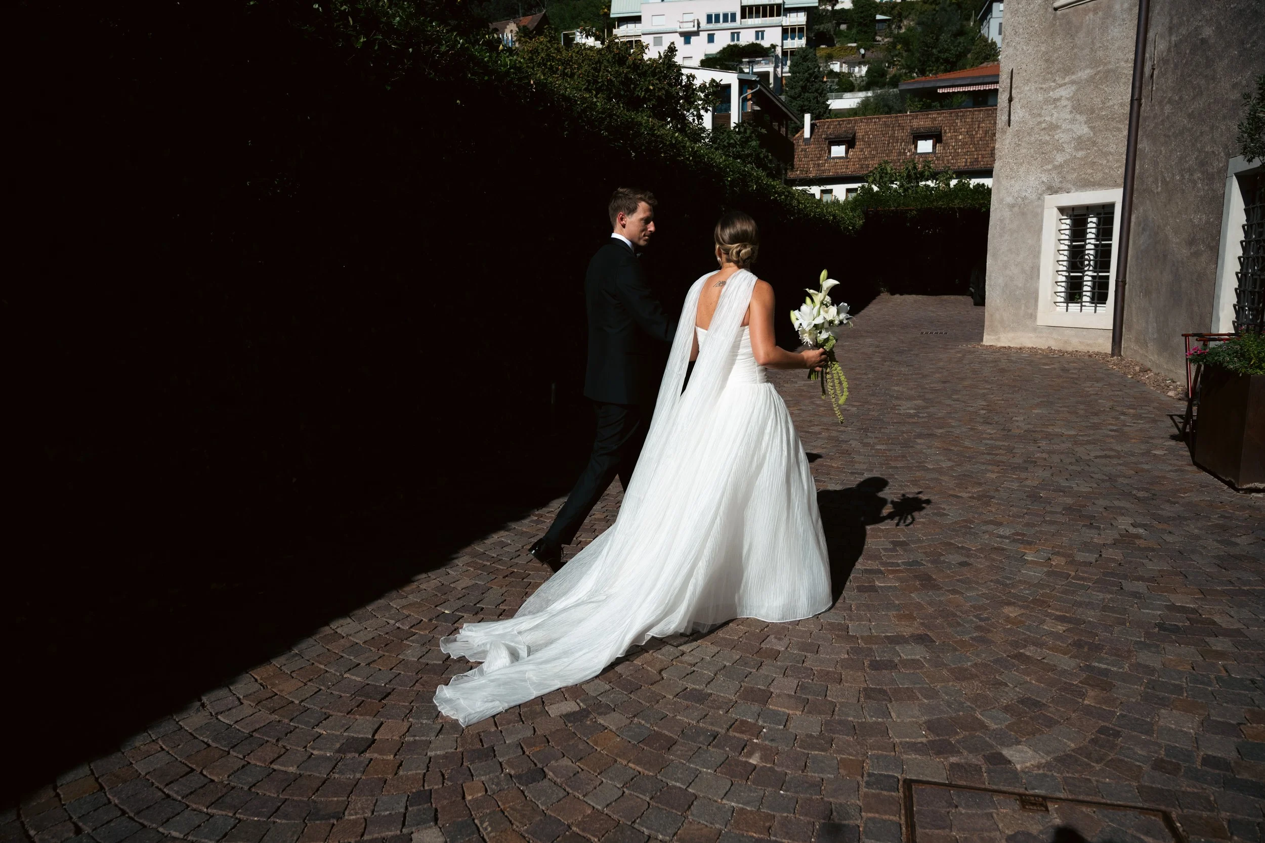 A bride and groom holding hands and walking outdoors on a cobblestone path during daytime, with the bride wearing a white wedding dress and holding a bouquet of flowers, and the groom in a black suit.