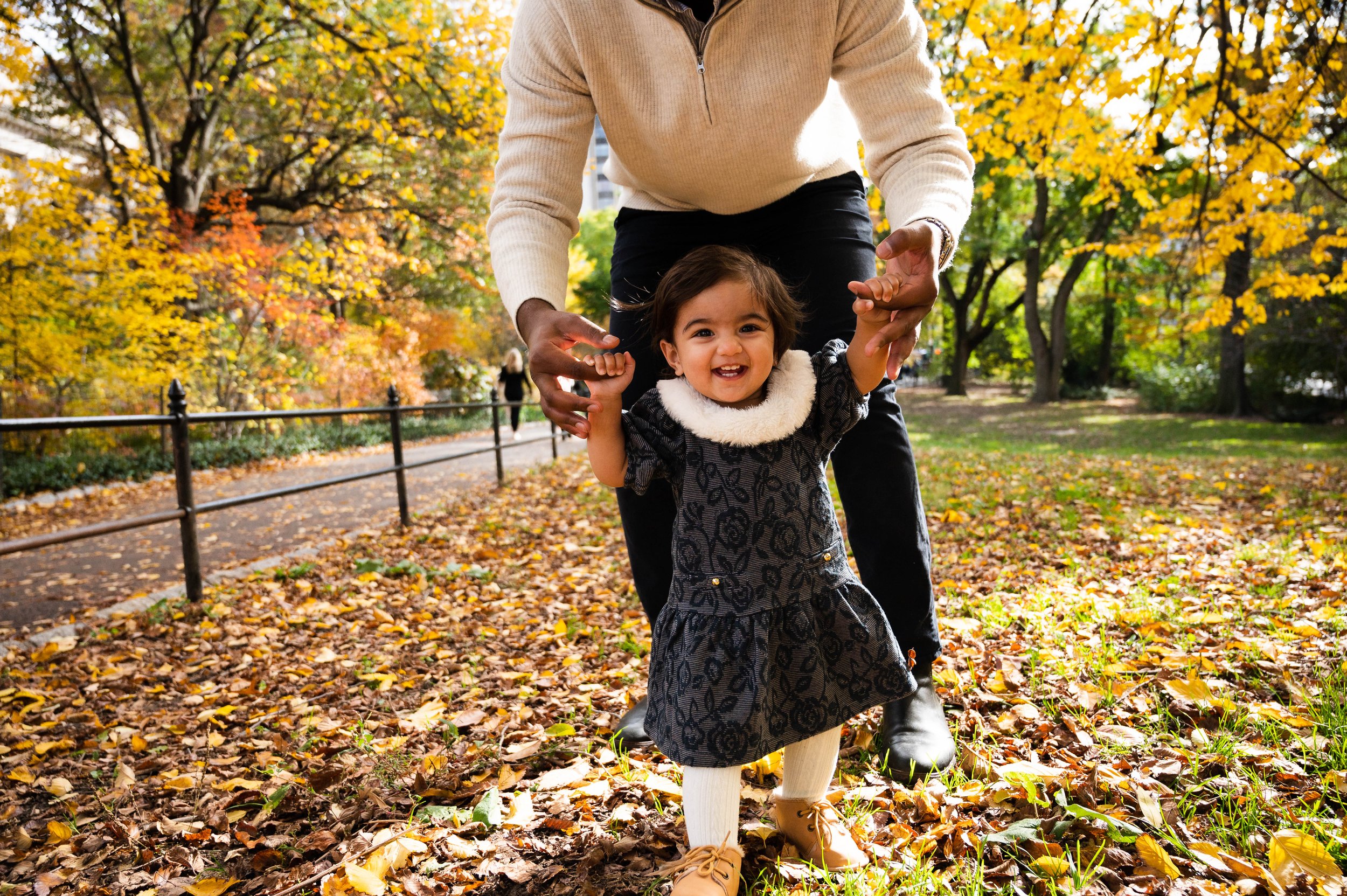 Alt text: Father holding his daughter's hands as she walks through Central Park during fall foliage season