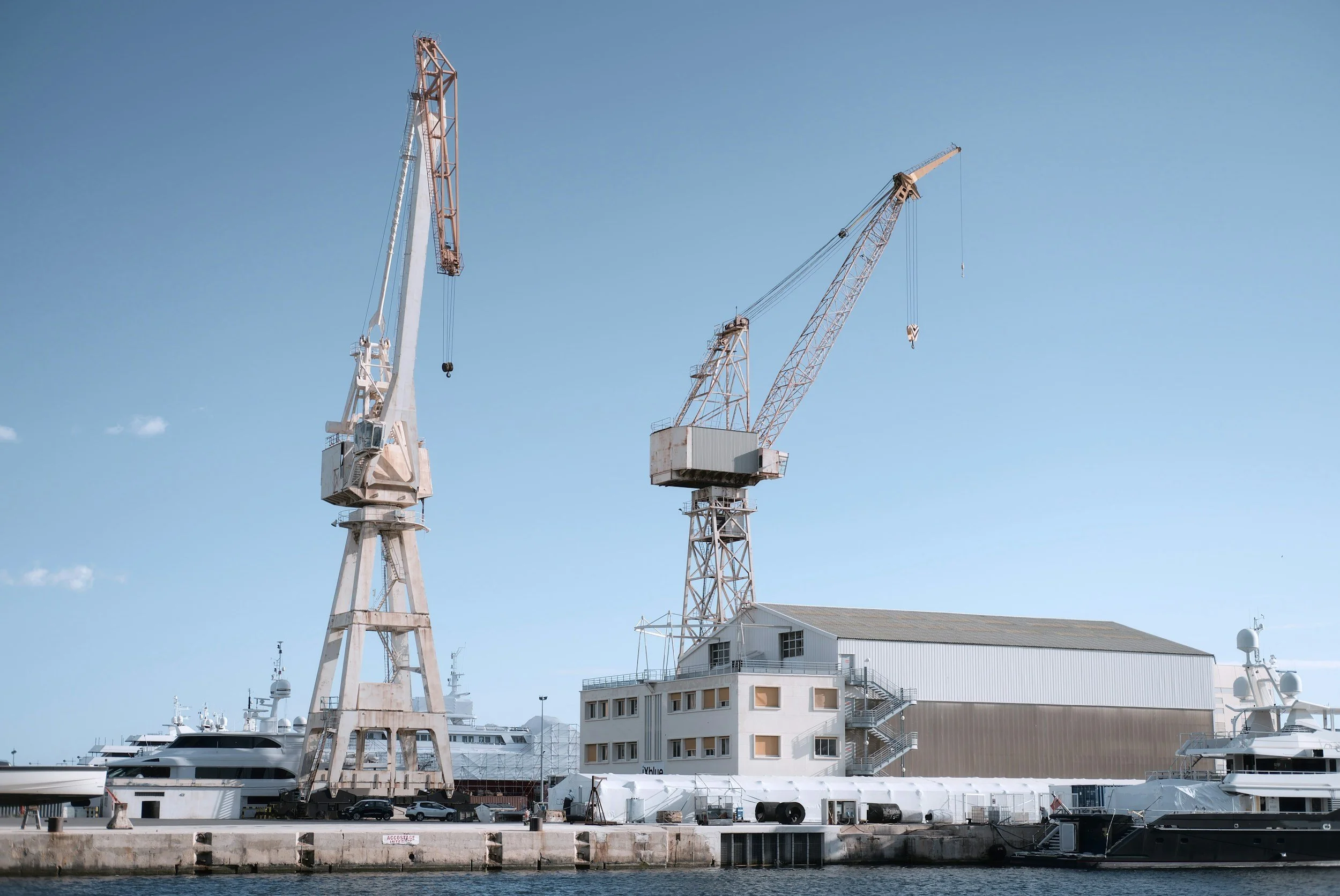 Large industrial cranes over a dock with yachts and boats, a warehouse building, and a blue sky with a few small clouds.