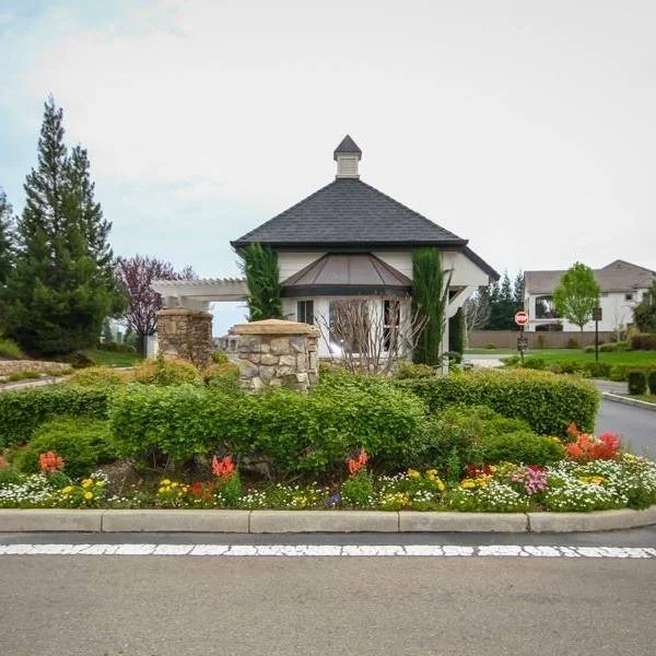 Small building with a dark gray roof and white walls, surrounded by bushes and colorful flowers, located near a street with a curb and sidewalk.