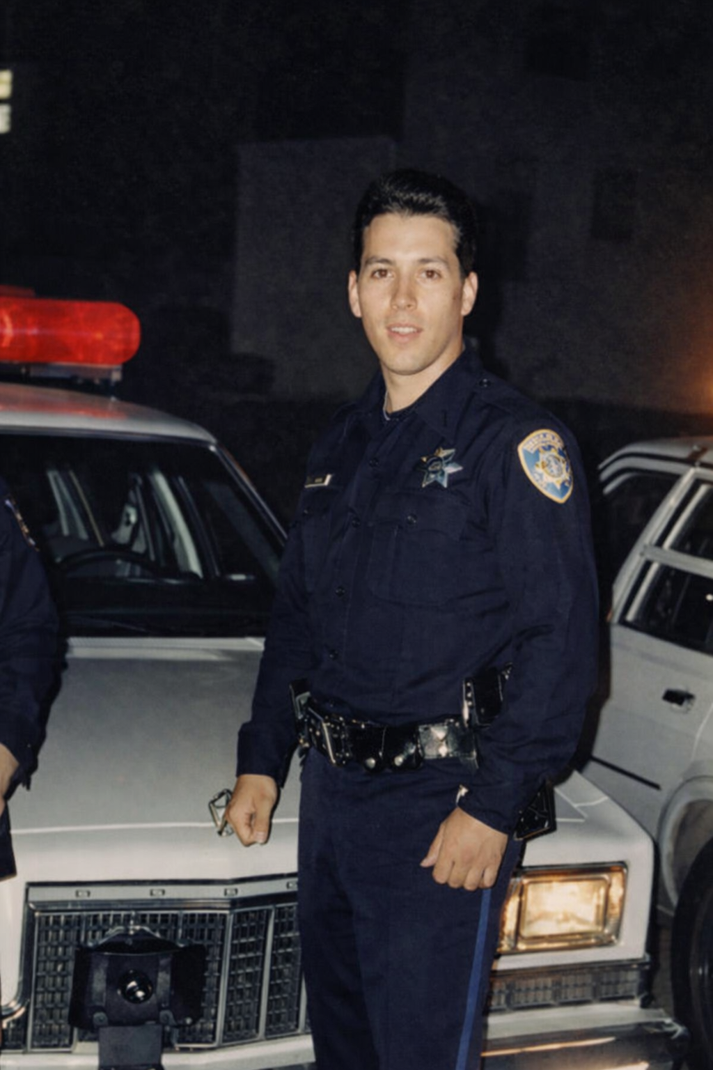 A young male police officer in uniform standing next to a police car at night, with a police badge visible on his shirt.