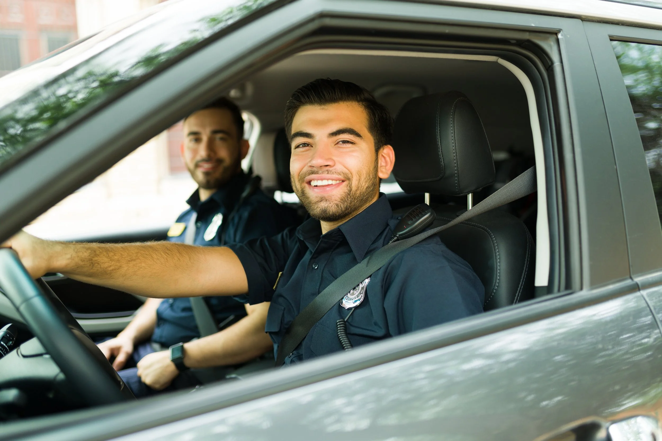 Two police officers sitting in a patrol car, smiling. The officer in front is leaning on the steering wheel, wearing a dark blue uniform with a badge and police radio. The officer in the back is also smiling, wearing a dark blue police uniform.