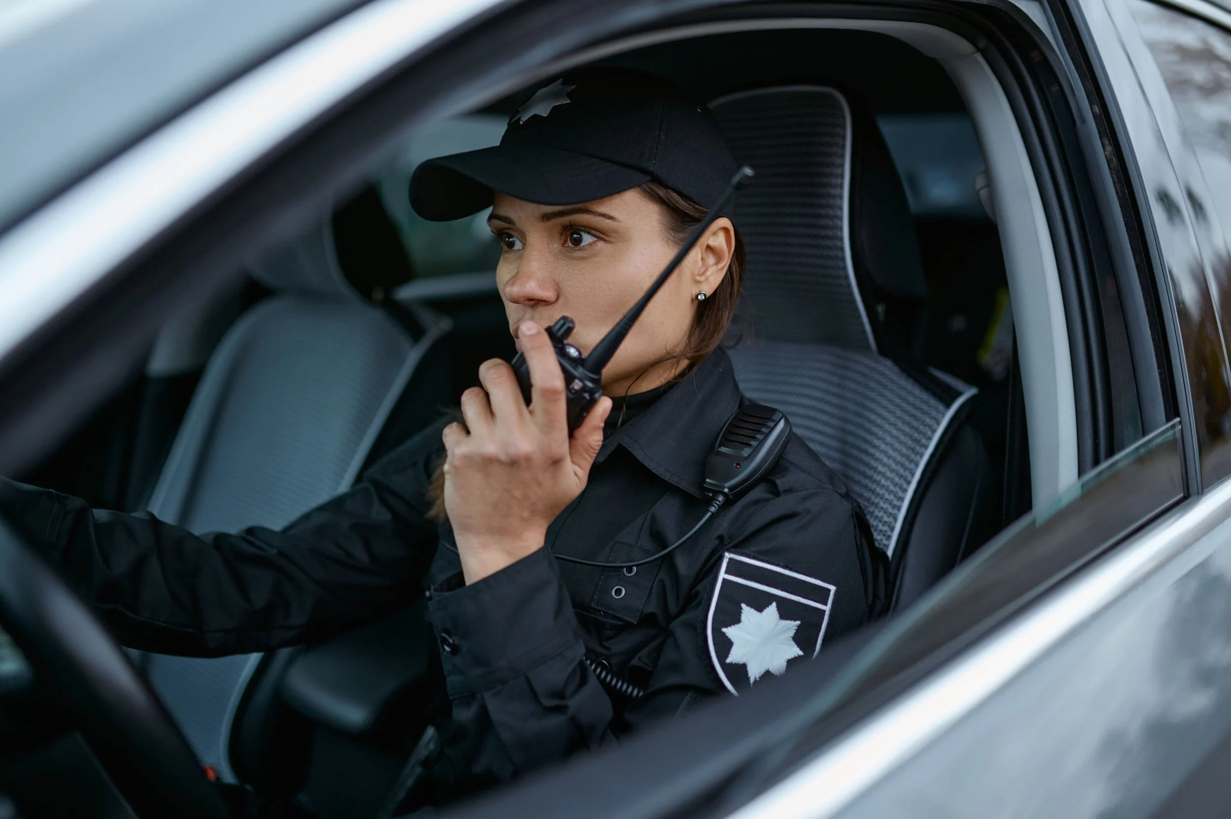 A female police officer sitting in the driver's seat of a police car, speaking into a radio microphone, wearing a black uniform and cap with a police badge on the sleeve.