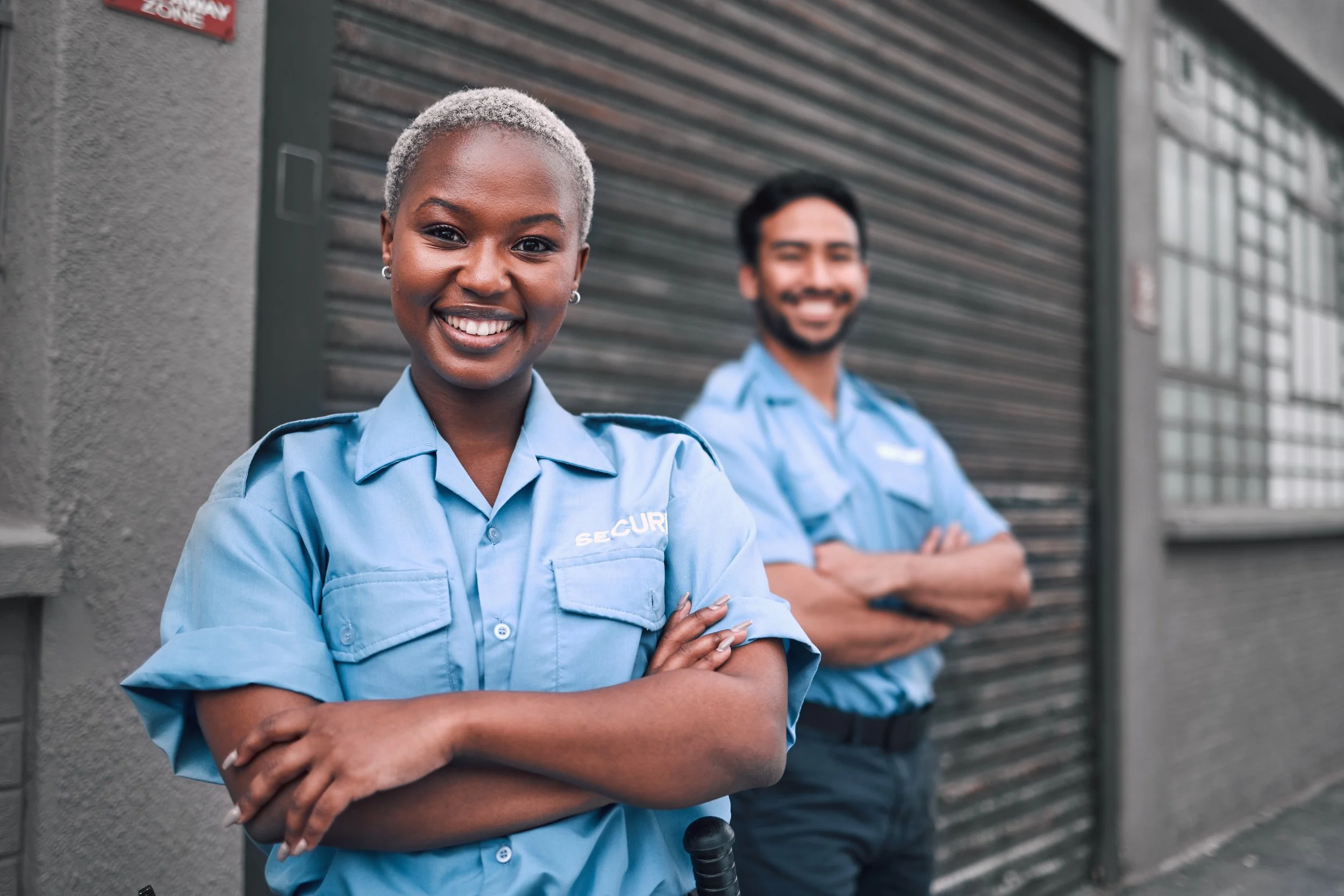Two security guards, a woman in the foreground with short gray hair and a man in the background, both smiling and wearing blue uniforms, standing outside near a building with a metal shutter.