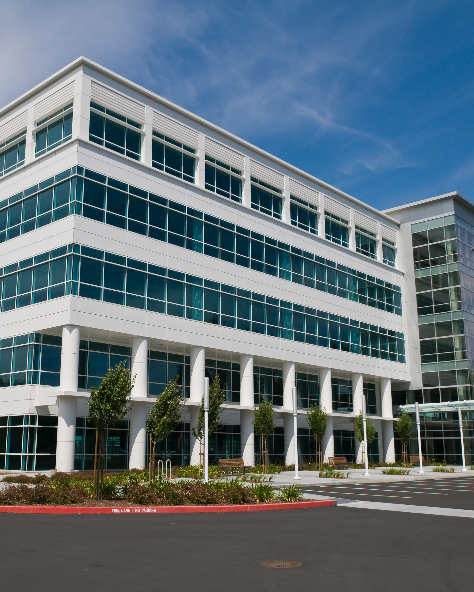 Modern multi-story office building with large glass windows under a blue sky, surrounded by trees and a parking lot.