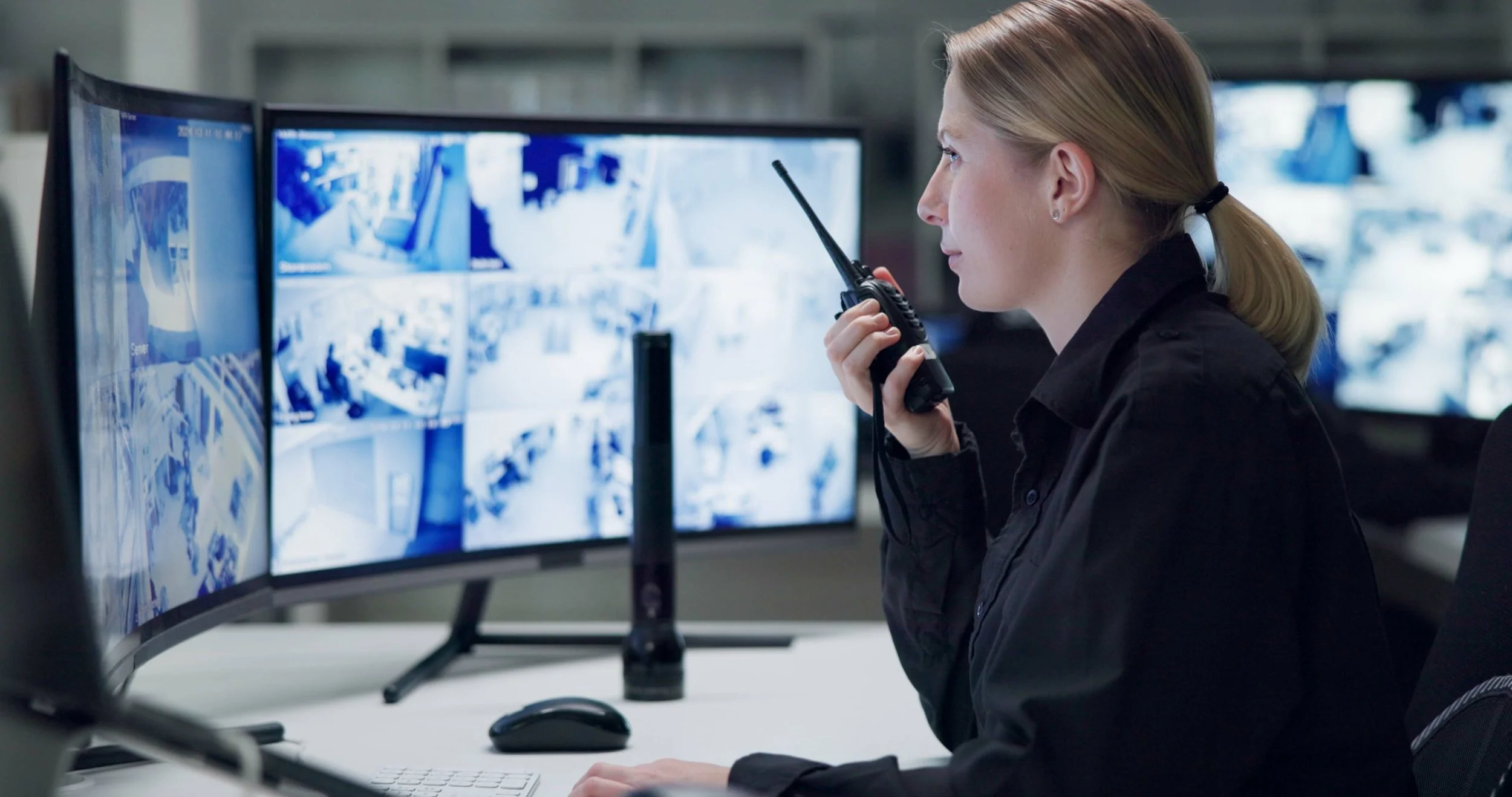 A female security officer in a control room monitoring multiple surveillance screens.