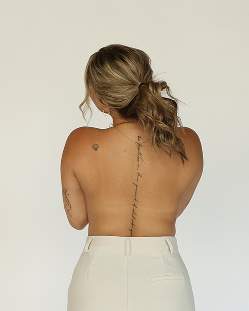 Back view of a woman with tattoos and blonde hair, wearing white pants, against a plain white background.
