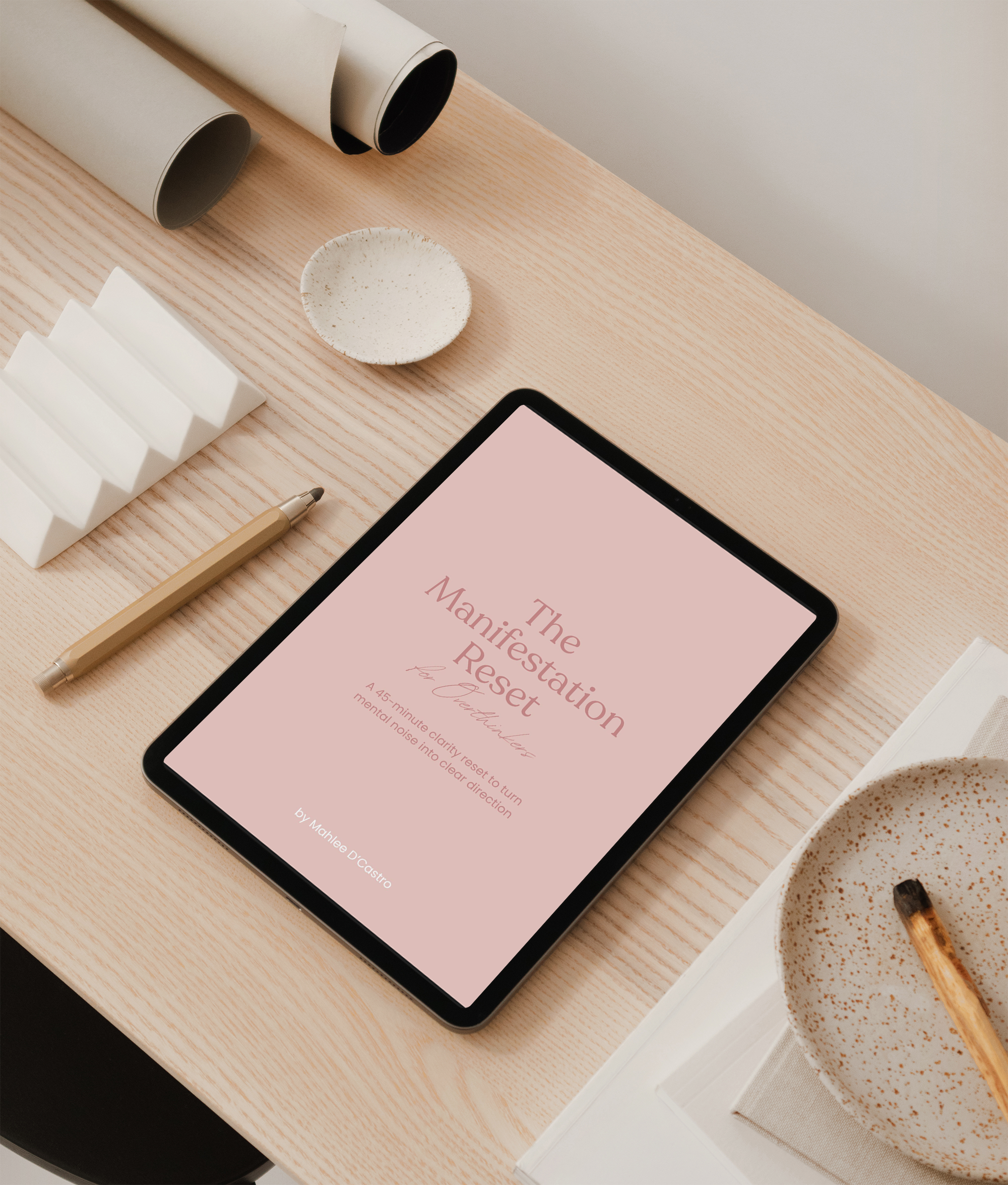 A digital tablet on a wooden desk displaying a book titled 'The Manifestation Reset' by Mahlee D'Castro, with a white bowl of coffee, a beige pen, a white layered notepad, and two rolled-up posters nearby.