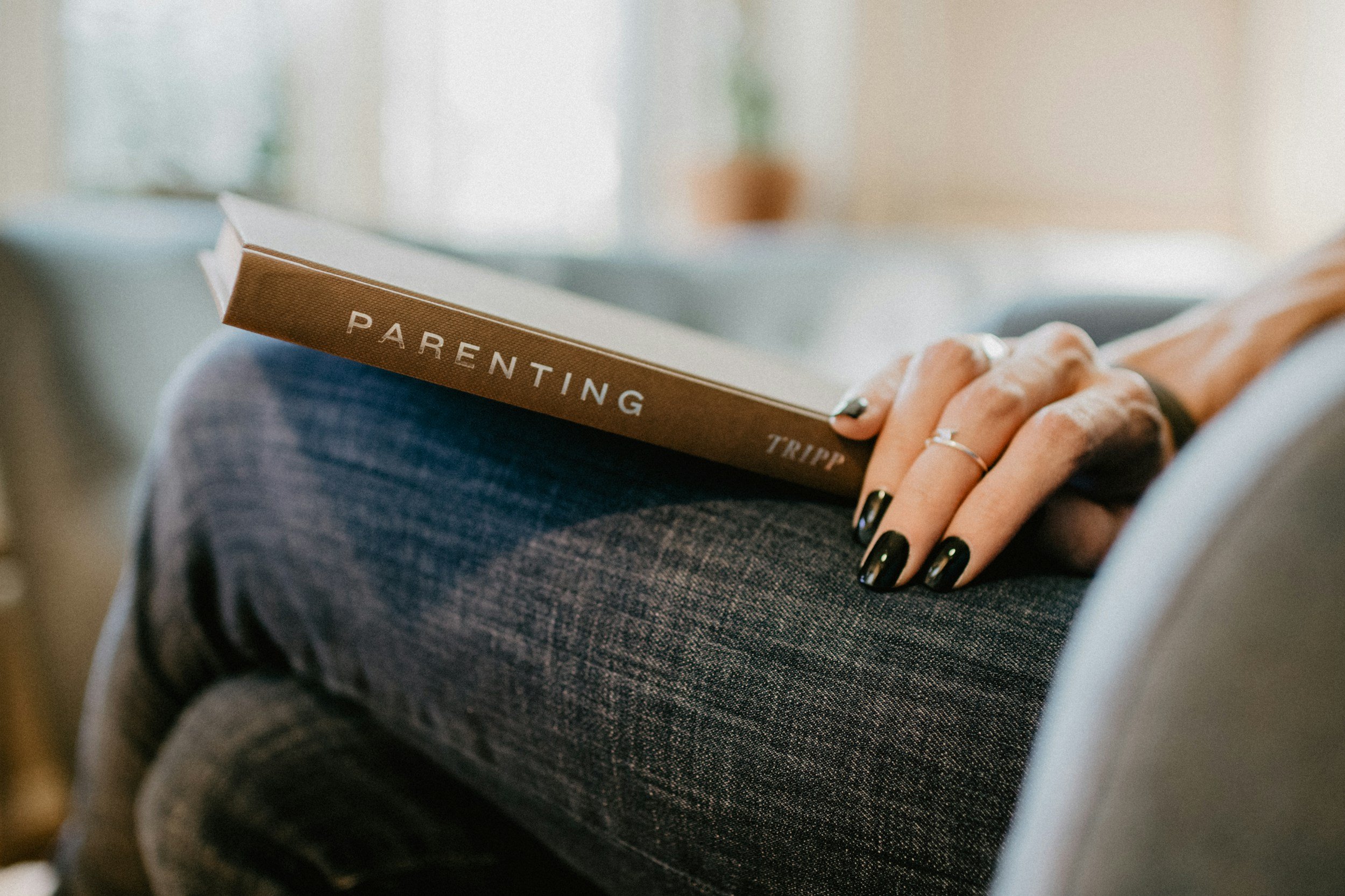 Person holding a book titled 'Parenting' in their lap, wearing dark jeans and painted nails in a parent coaching session.