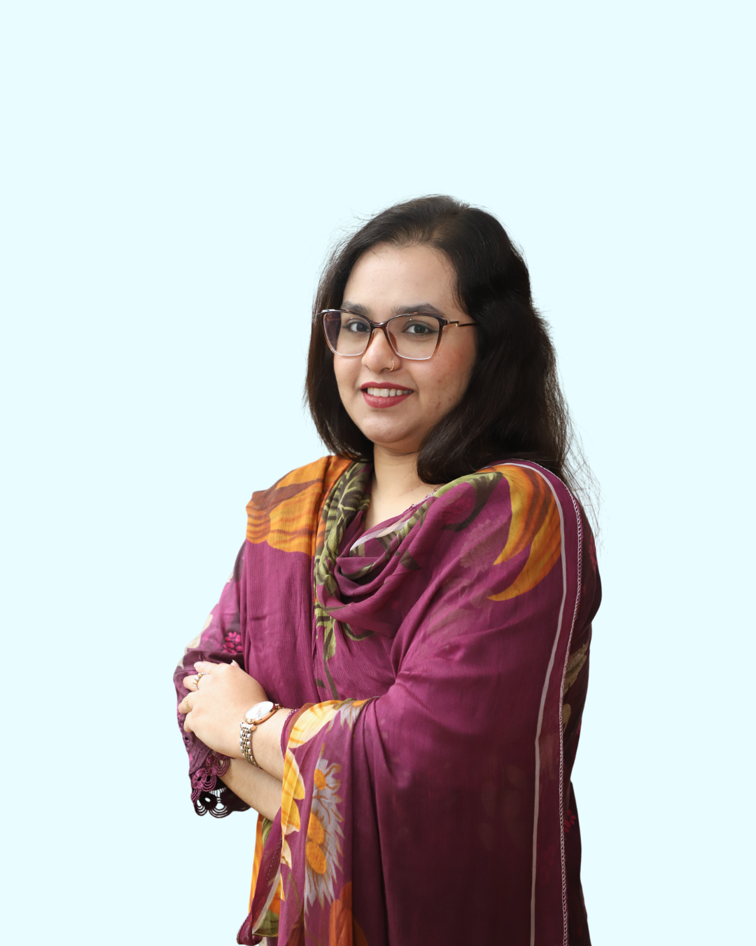 A woman with glasses, dark hair, and a nose piercing wearing a colorful floral-patterned sari, smiling with her arms crossed against a light blue background.