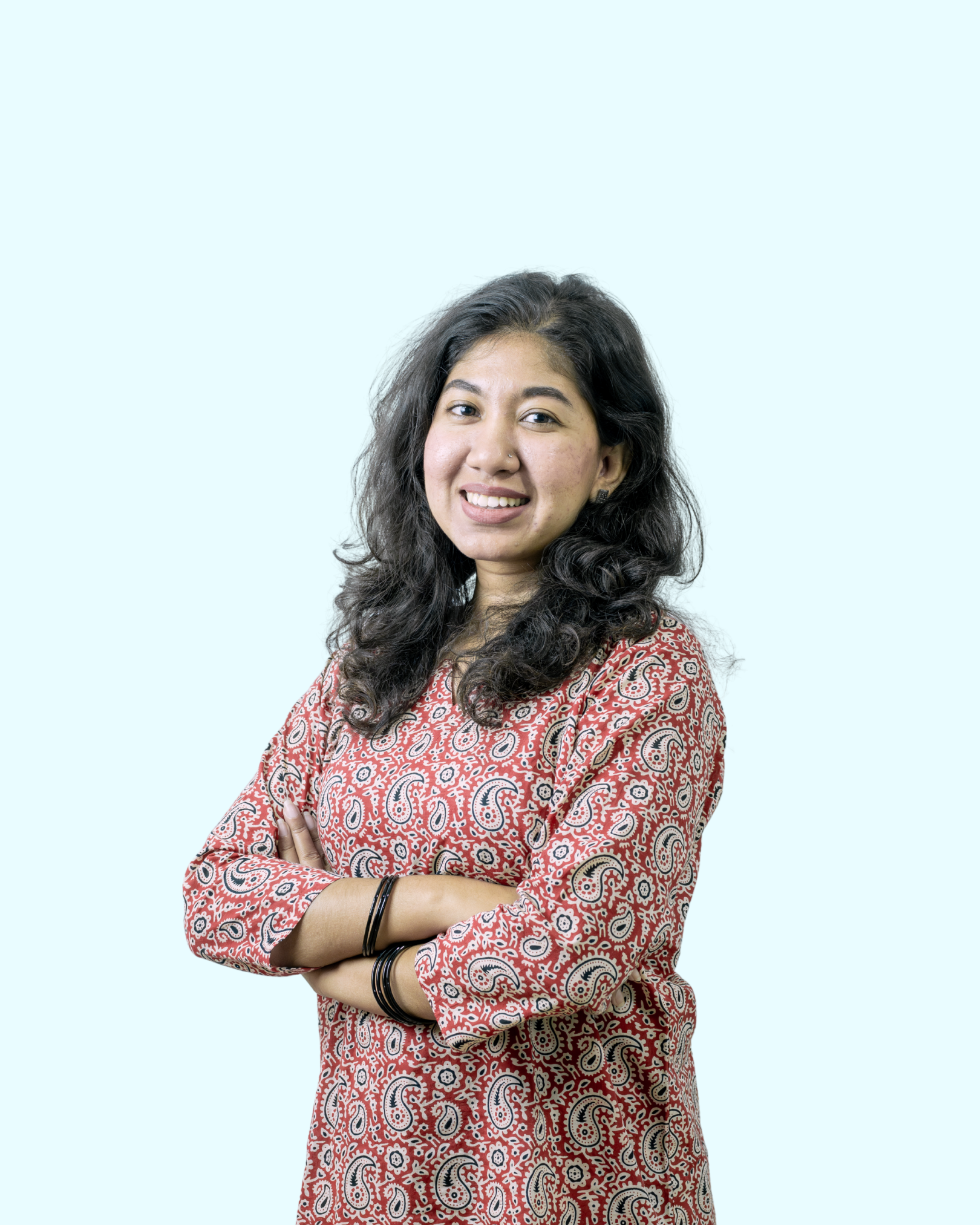 A young woman with long curly dark hair, wearing a patterned red and beige top, smiling with arms crossed against a light blue background.